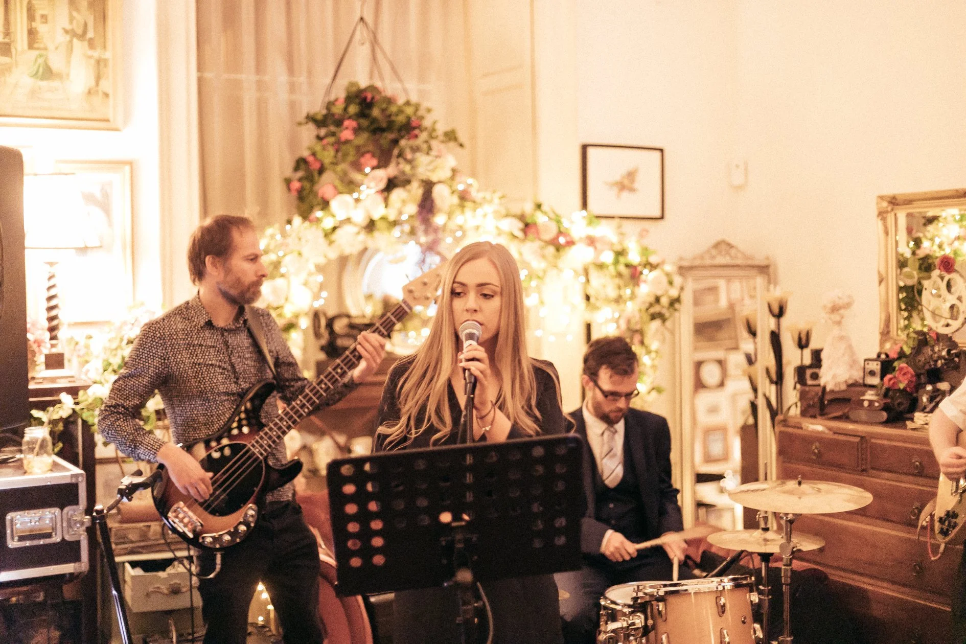A band performing at an indoor event with floral decorations and framed pictures, including a female singer, a guitarist, and a drummer.