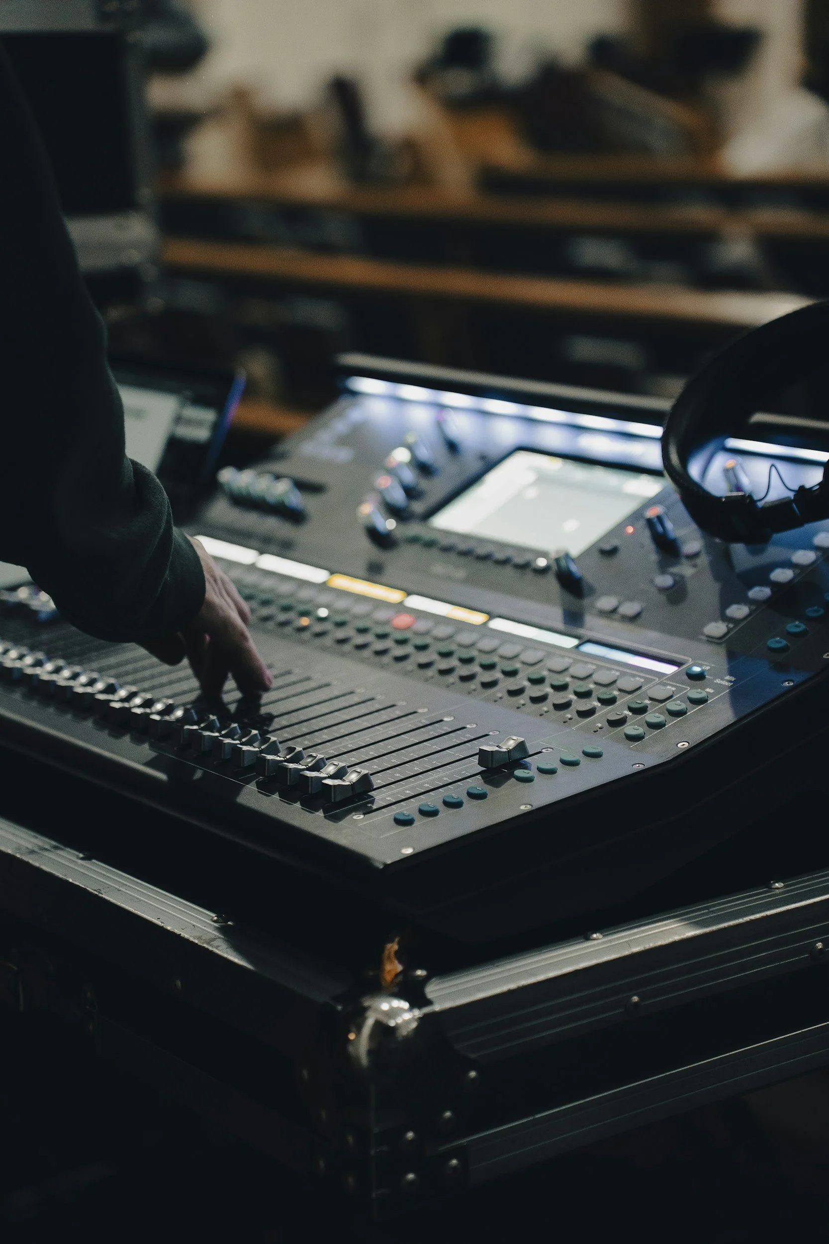 A person controlling a DJ mixing console with numerous knobs and sliders in a dark environment, with shelves of vinyl records in the background.