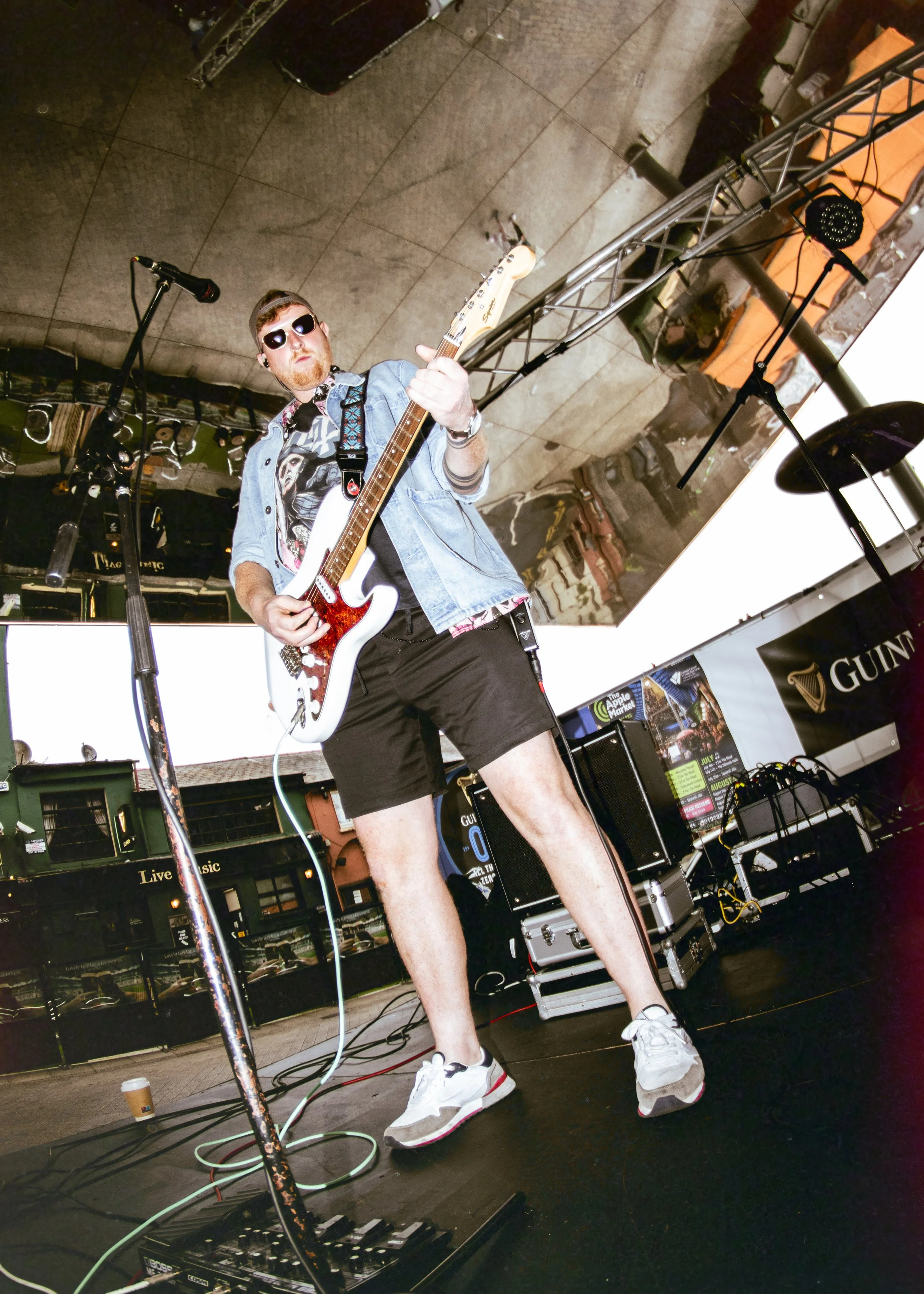 Male musician playing guitar on stage, wearing sunglasses, a denim shirt over a graphic t-shirt, black shorts, and sneakers, with stage equipment and background behind him.
