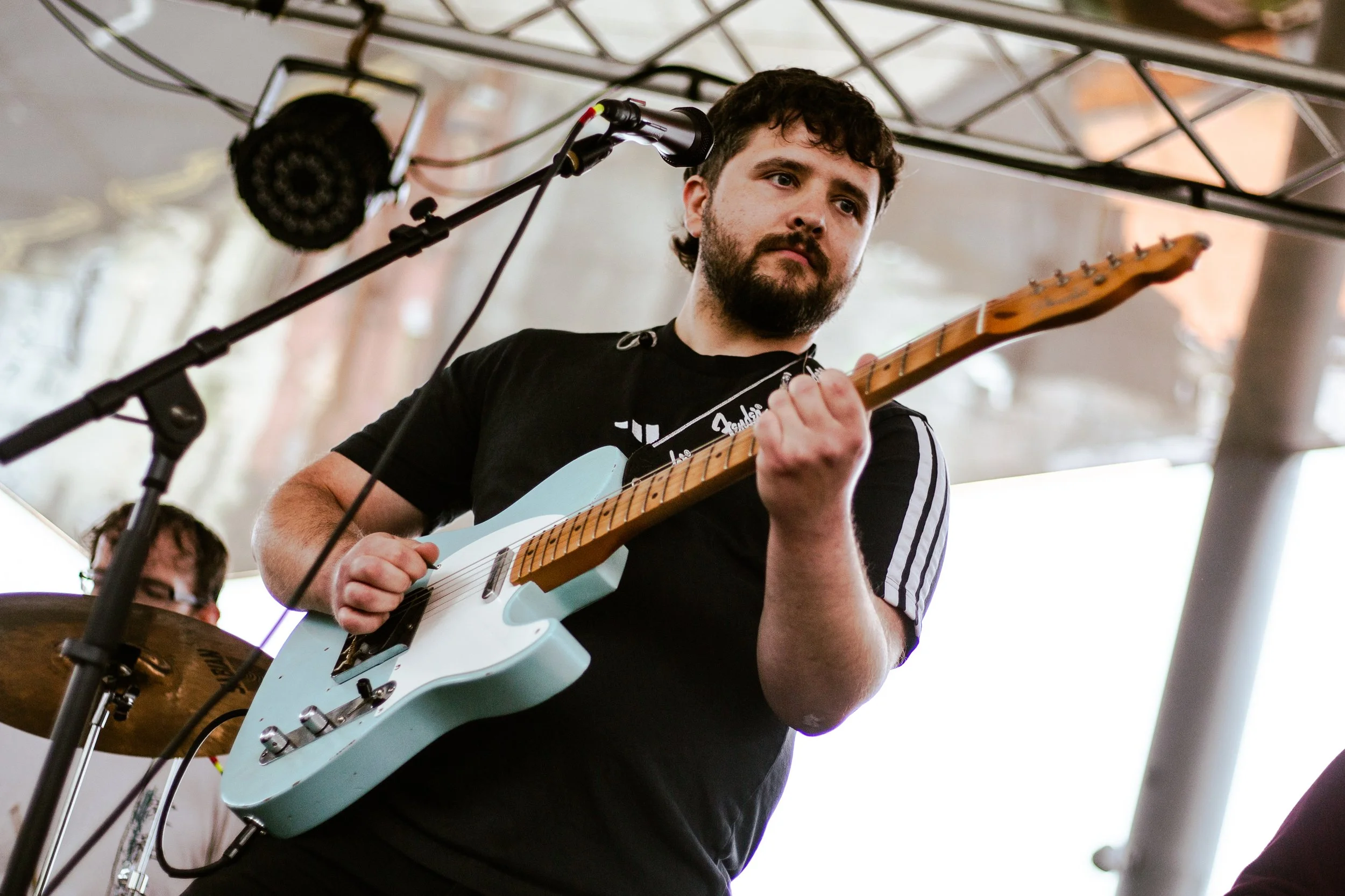 A man with dark curly hair and a beard playing an electric guitar during a live performance, with a microphone in front of him and a drummer partially visible in the background.