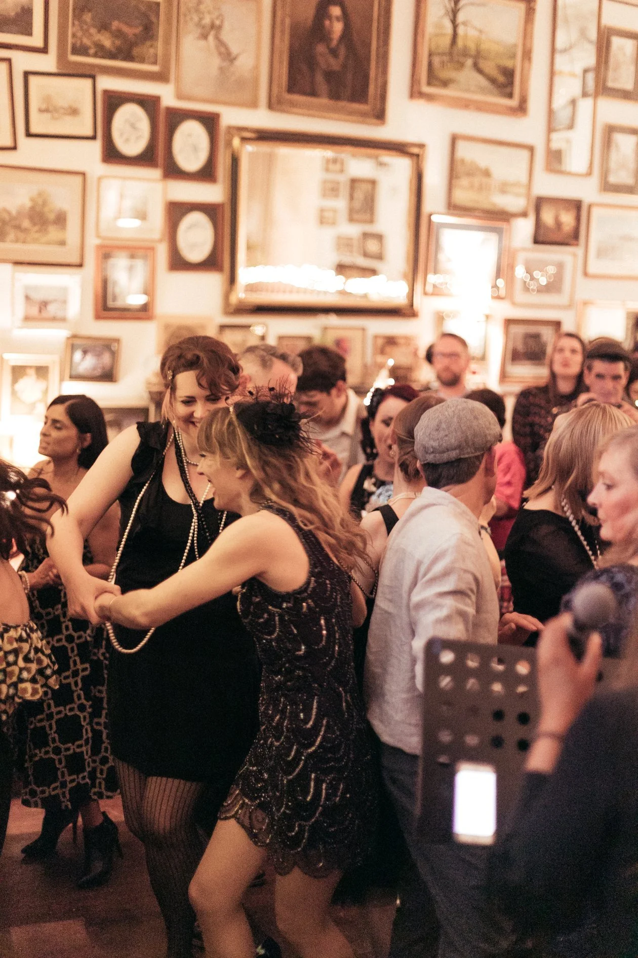 People dancing at a party or social gathering in a warmly lit room decorated with numerous framed pictures and artwork on the walls.
