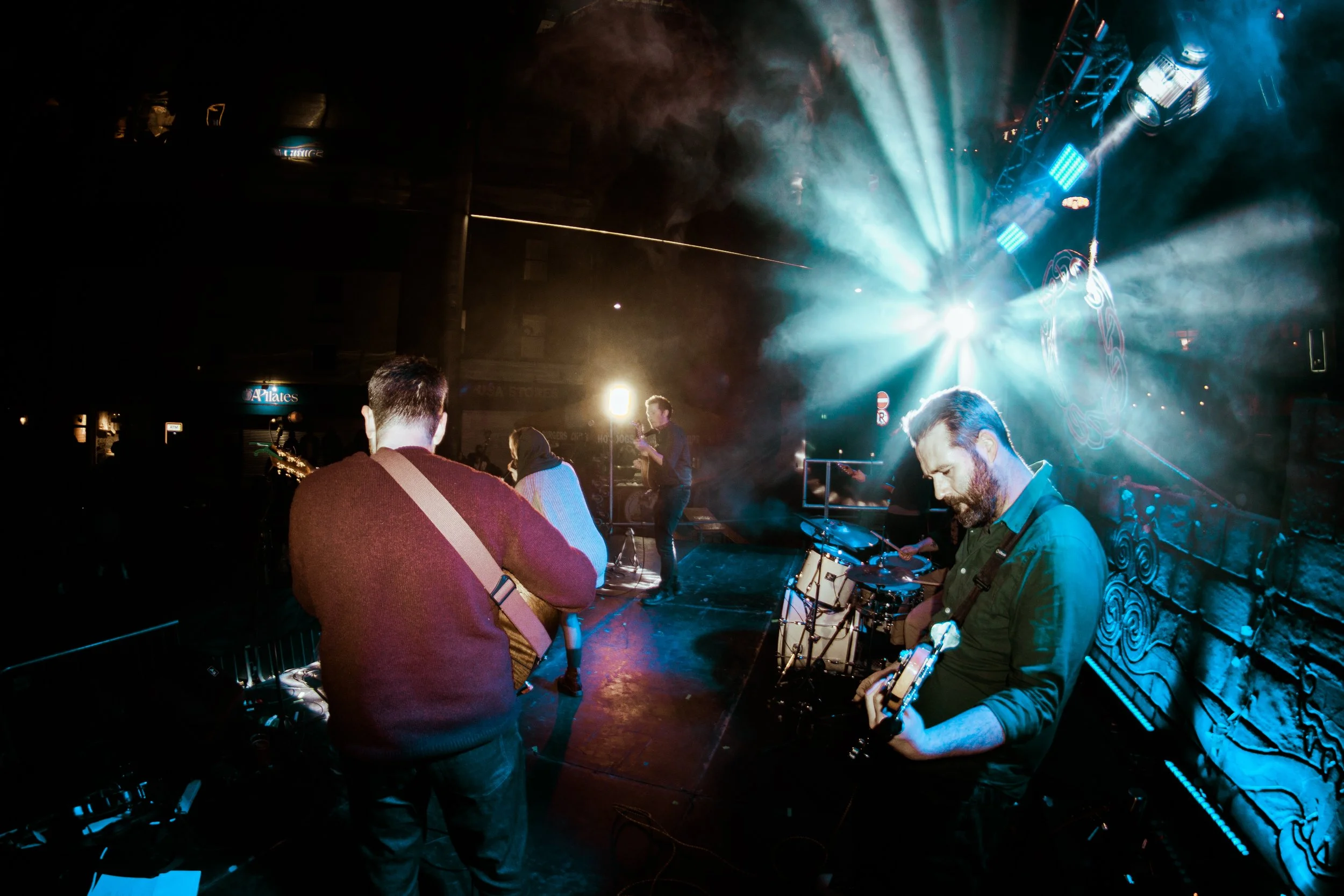 Musicians performing on stage at a concert with blue and white lights, smoke effects, and an audience watching.