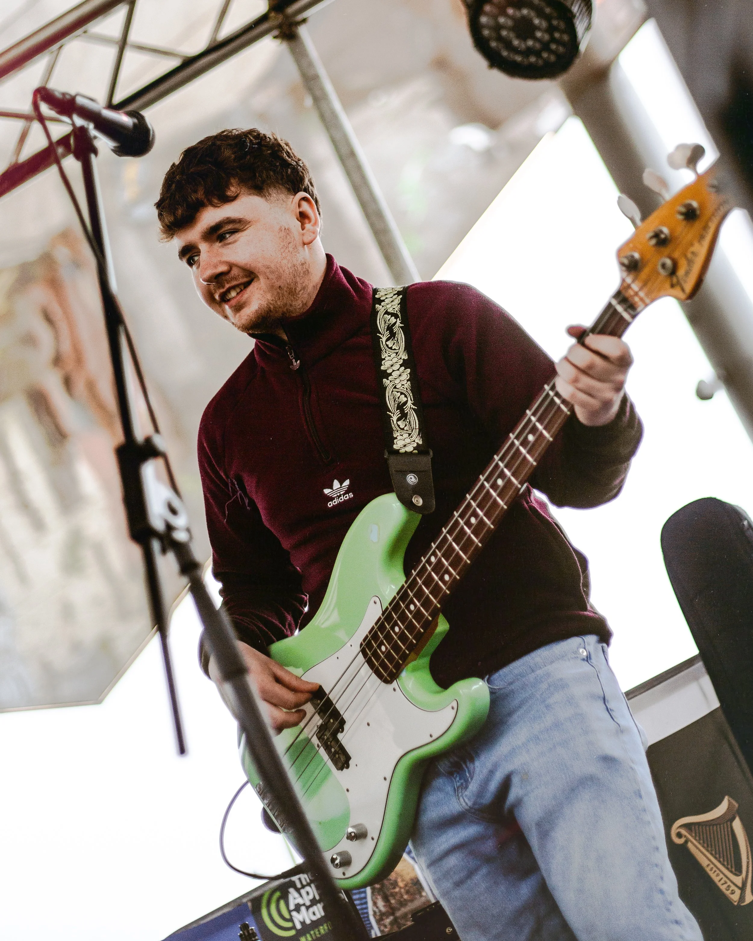 A young man playing an electric bass guitar with a green body and white pickguard, wearing a maroon jacket and light blue jeans, standing in front of a microphone and a sound equipment setup.