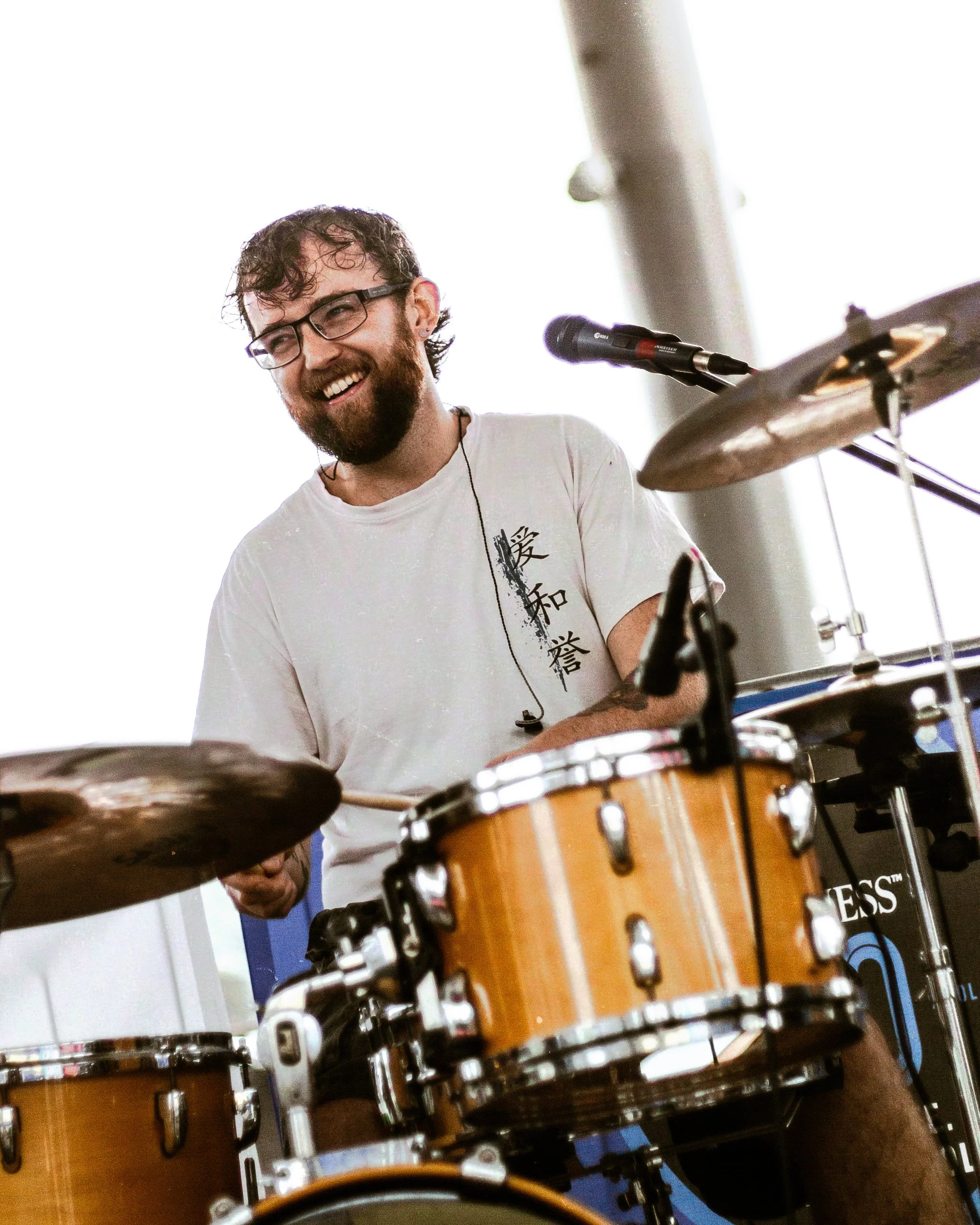 A man with glasses and a beard smiling while playing drums during a performance.