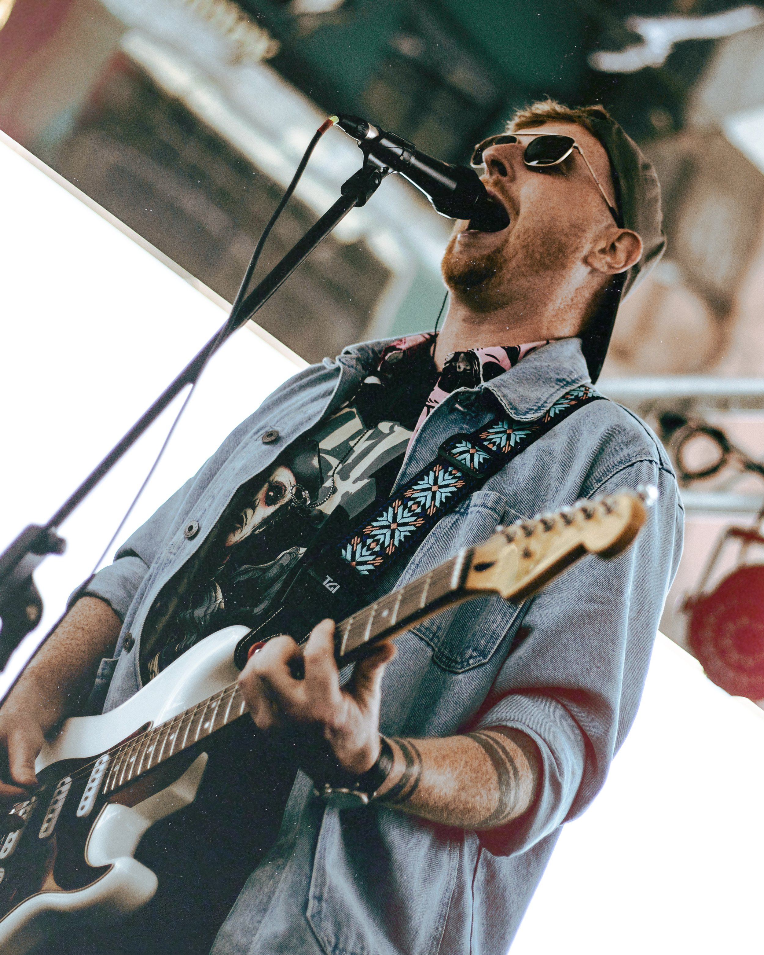 A man wearing sunglasses, a baseball cap, and a denim jacket playing a white electric guitar and singing into a microphone.