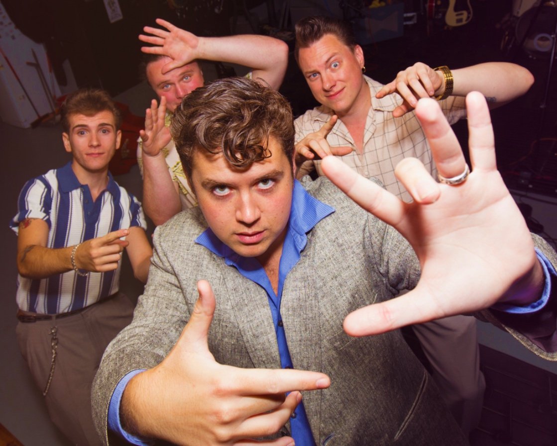 Four young men posing playfully for the camera, with one in the foreground making a hand gesture and two others in the background making playful expressions.