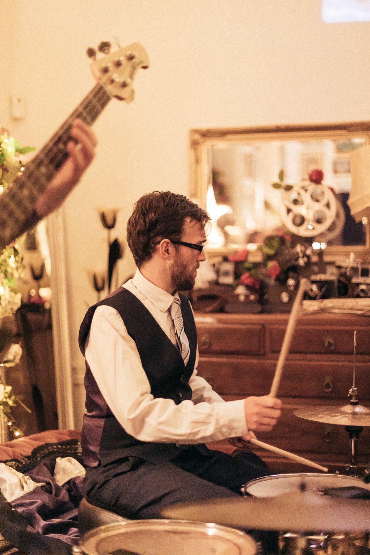 A man playing drums. He has curly hair, glasses, and is wearing a white shirt with a black vest and dark pants, seated in a room with warm lighting, a wooden dresser, mirror, and decorative items.