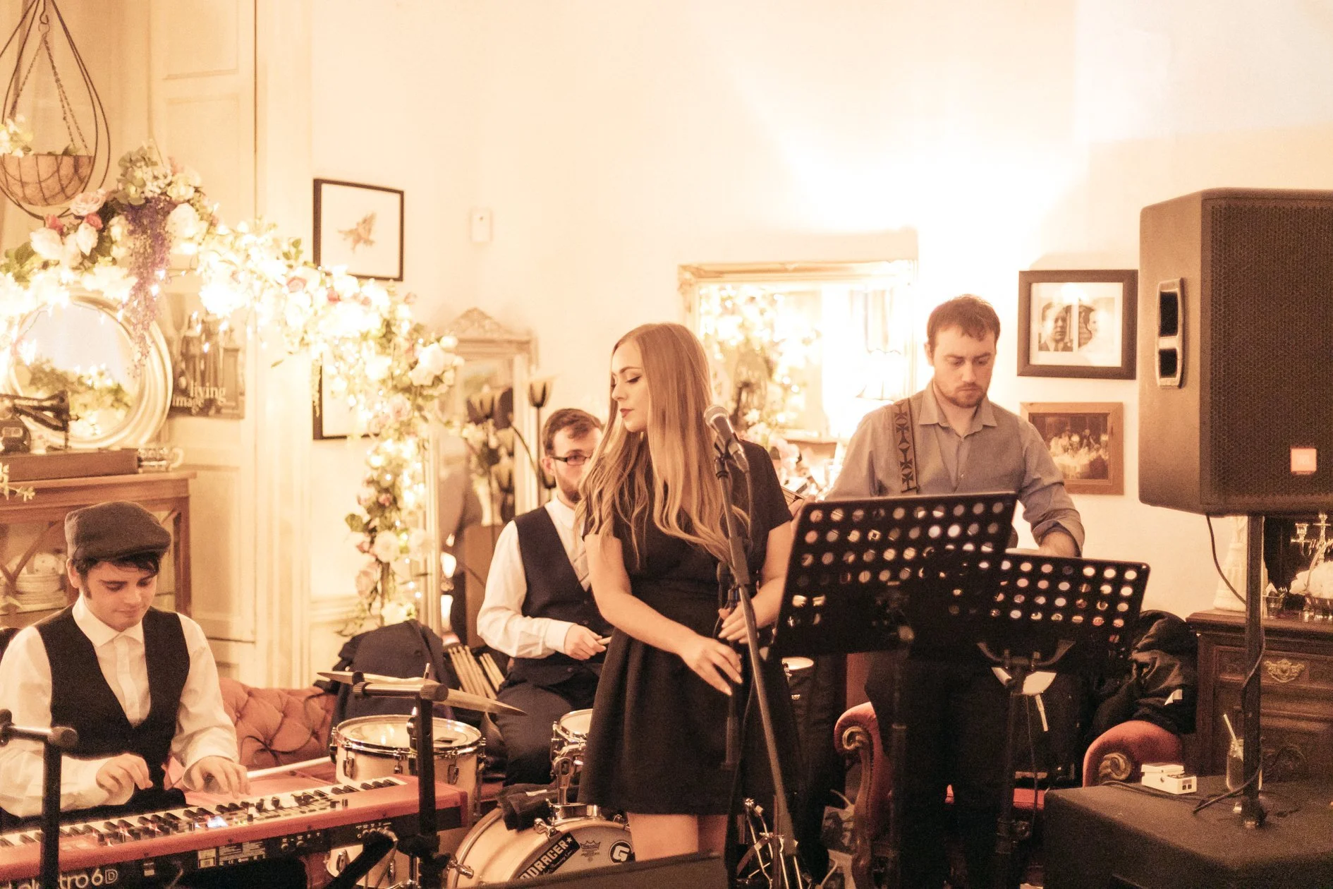 A group of musicians performing indoors, including a young woman singing into a microphone, a keyboard player, and a drummer among other band members, with floral decorations and framed photos on the wall.