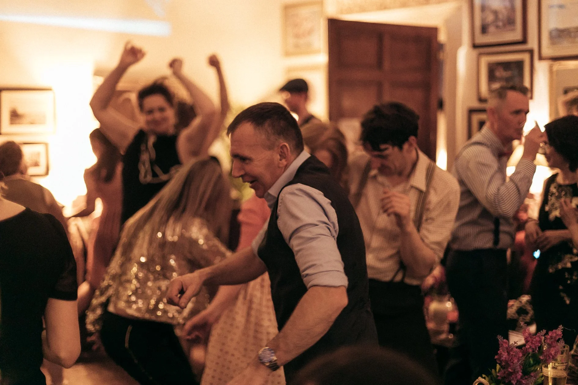People dancing and socializing at a lively indoor party or celebration with warm lighting and framed pictures on the walls.