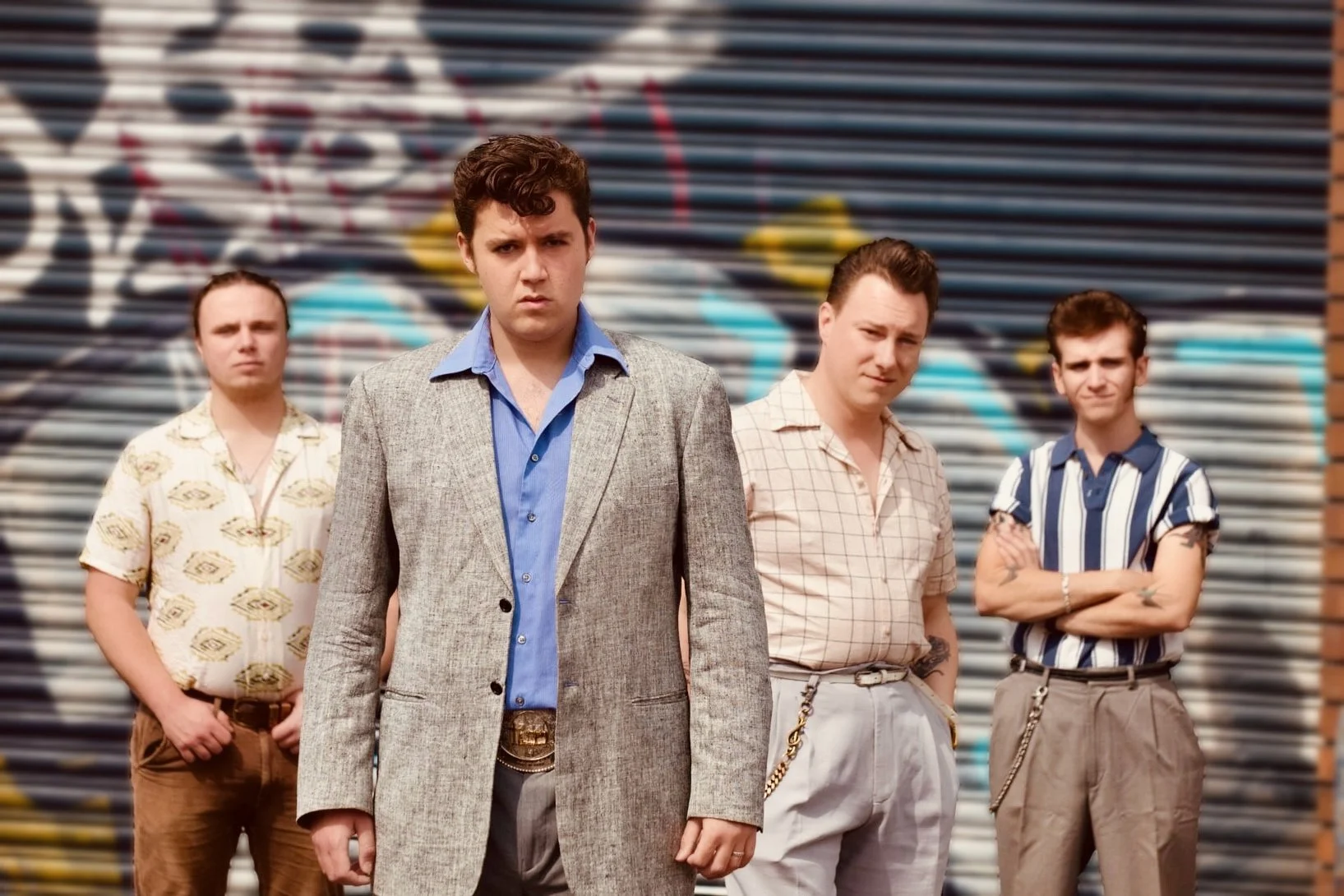 Four young men standing in front of a graffiti-covered metal roller shutter, dressed in vintage 1980s attire, with serious expressions.