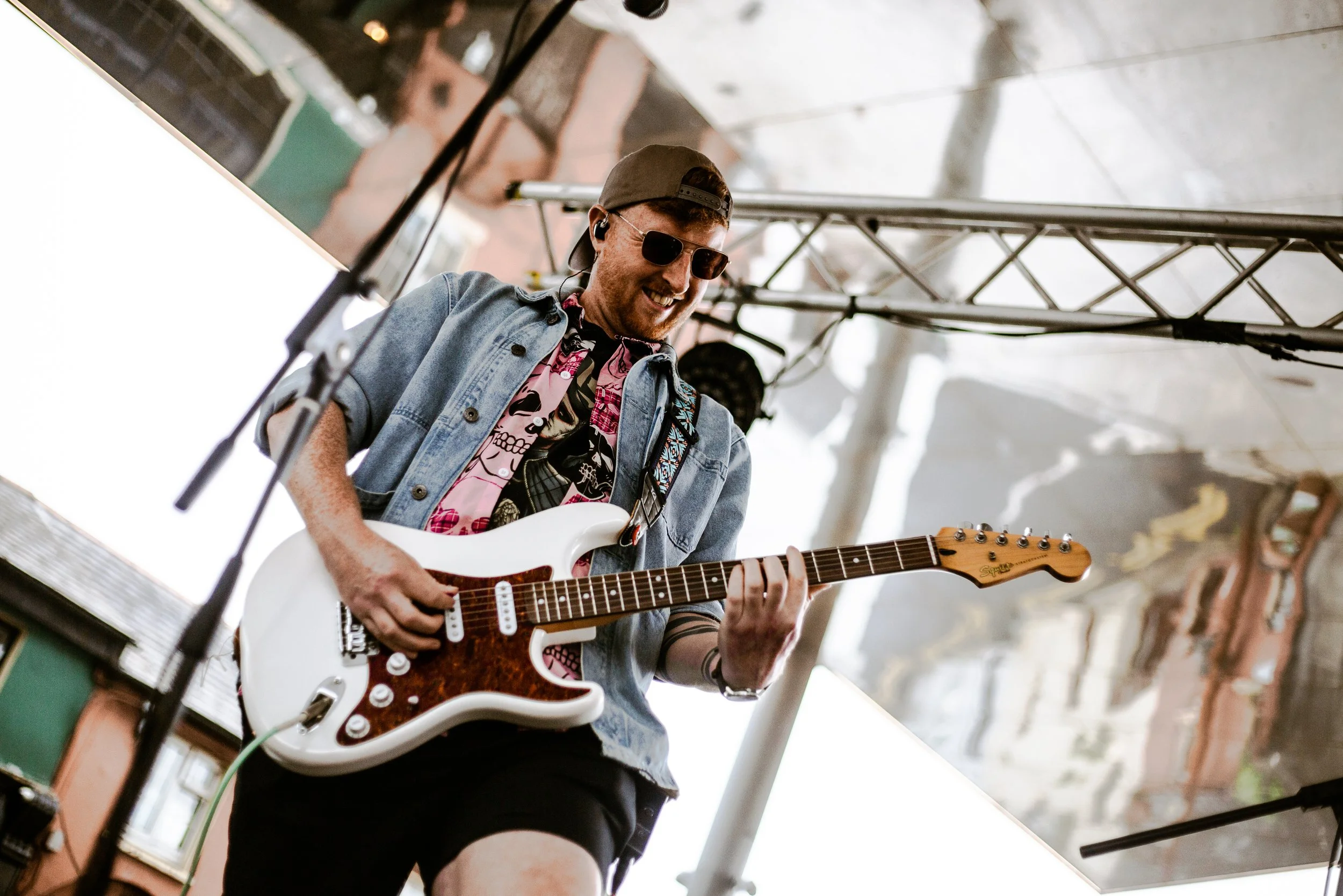 A young man wearing sunglasses, a backward cap, and a colorful shirt under a denim jacket plays an electric guitar on a stage, smiling and enjoying the music. There are musical equipment and a pole in the foreground.