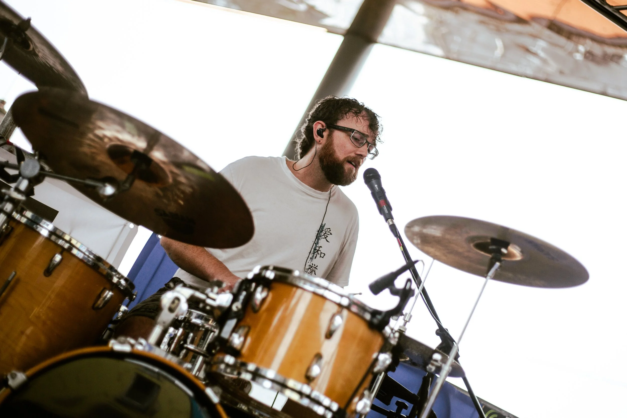 A man with glasses and a beard is playing drums on stage, wearing an earphone, with a microphone in front of his mouth.