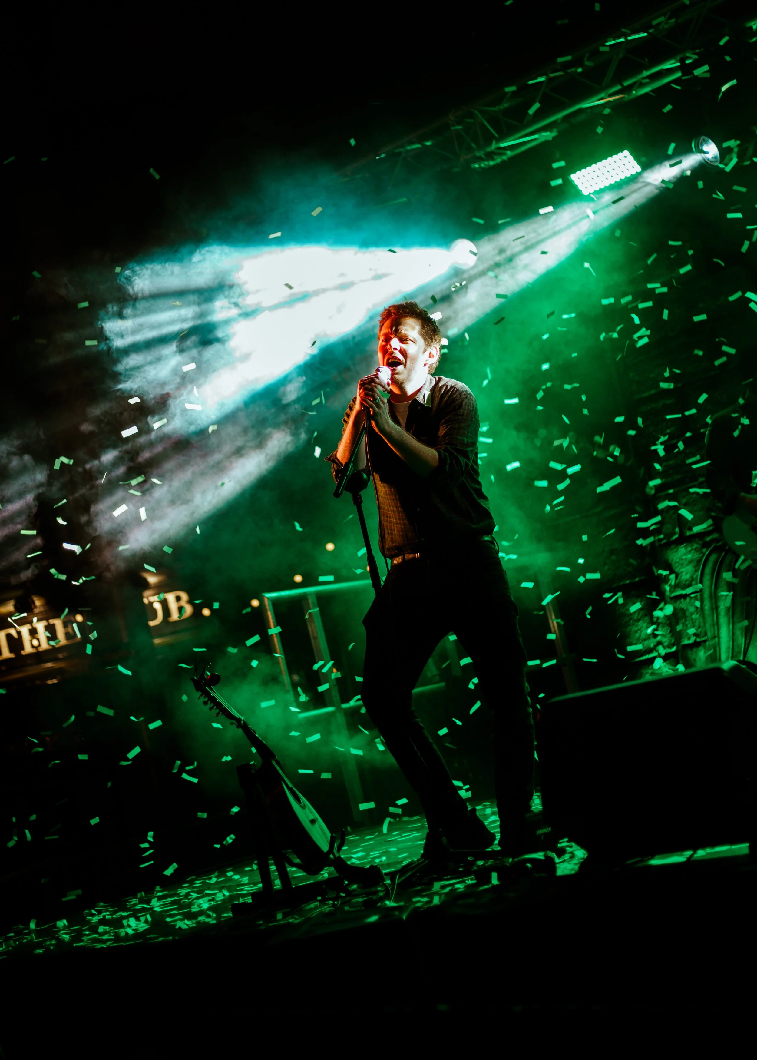 A male singer on stage holding a microphone, illuminated by dramatic green and white stage lighting, with confetti falling around him.