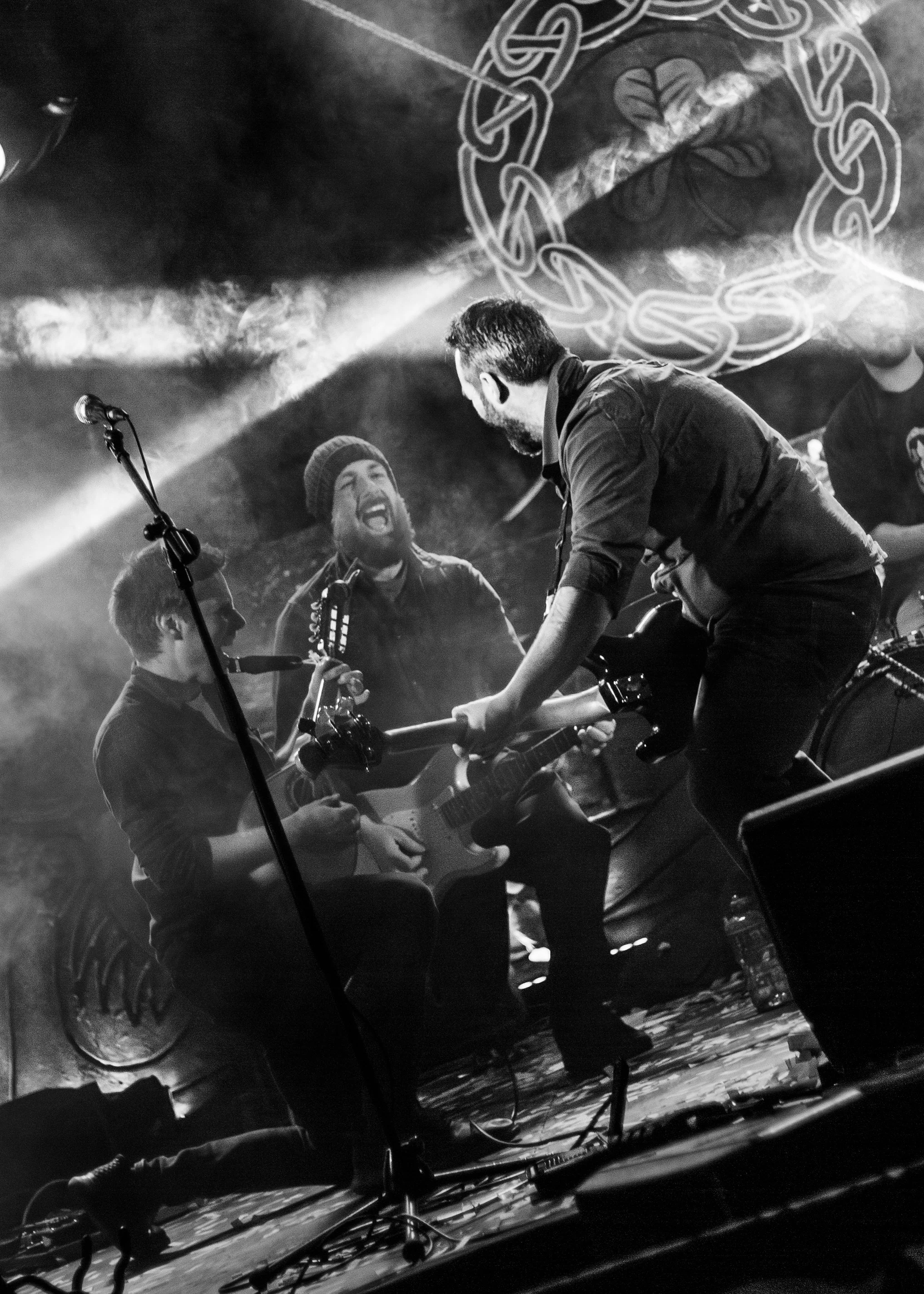 Black and white photo of four musicians performing on stage, with one playing a guitar, another singing, and the others playing instruments, with a decorative backdrop featuring intertwined circular designs and floral motifs.