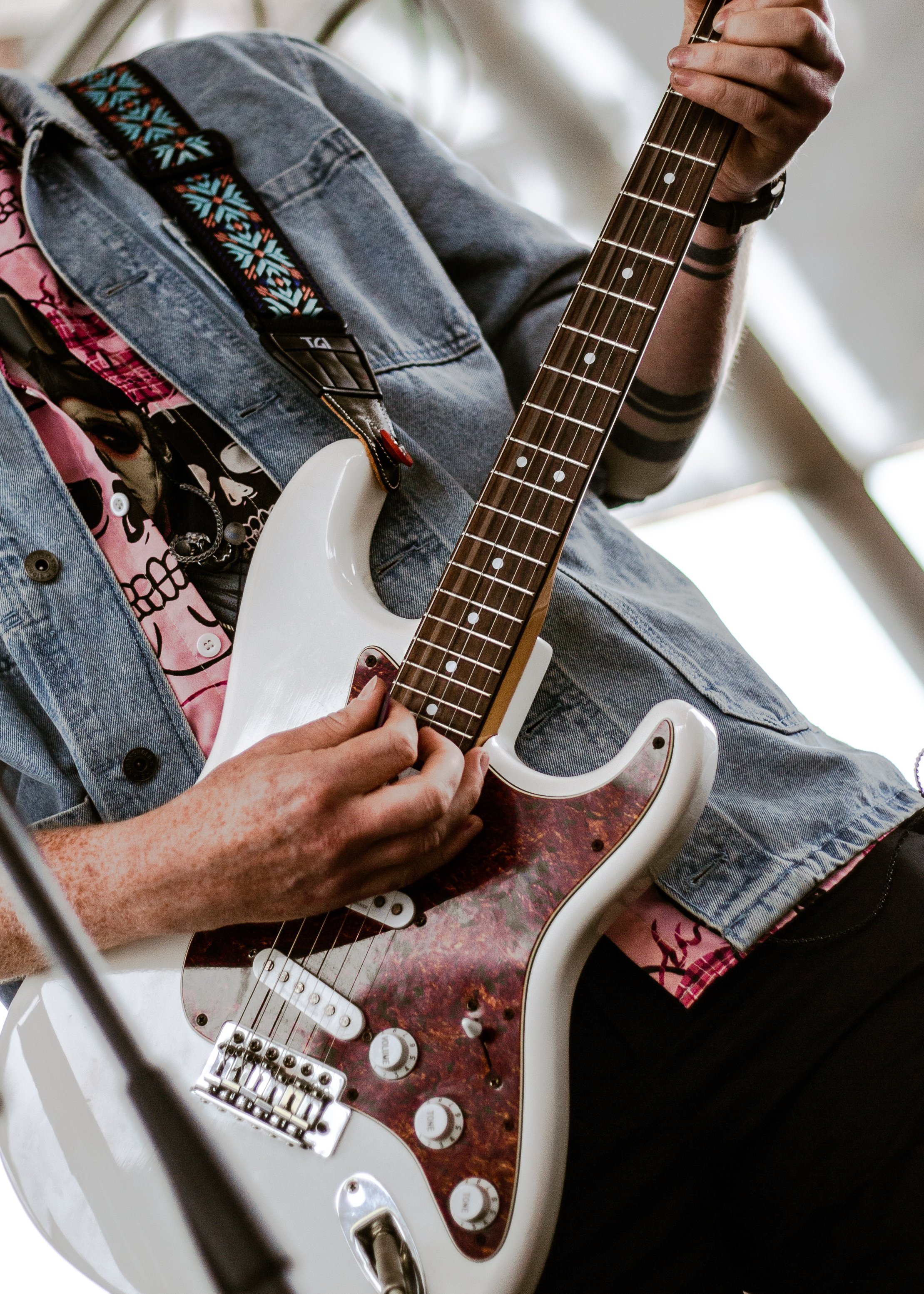 Person playing an electric guitar with a sunburst pickguard, wearing a denim jacket over a pink skull-print shirt, with a black wristband and tattoos on their arm.