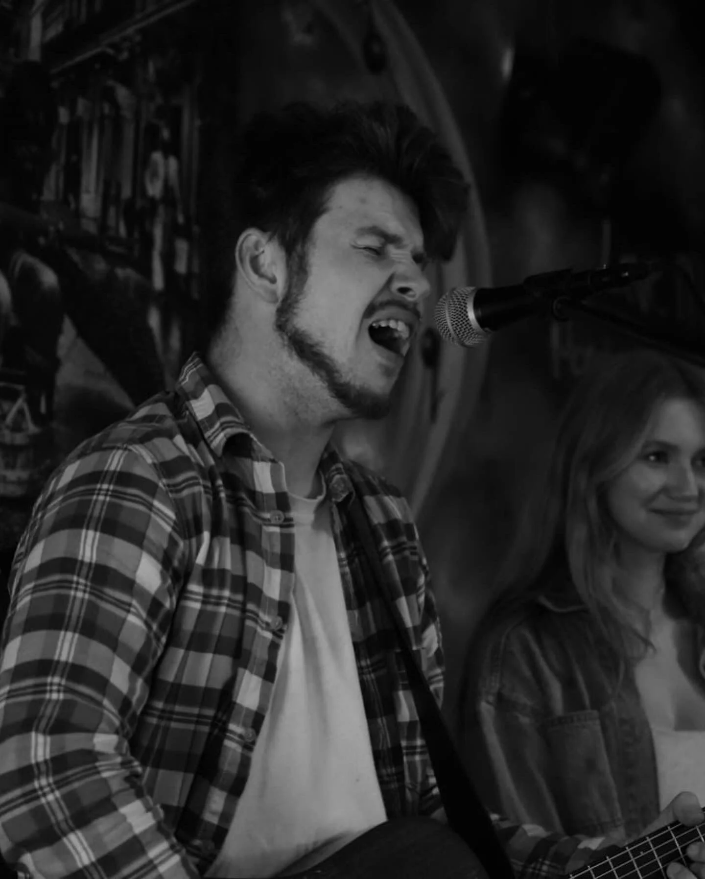 A young man singing passionately into a microphone while playing an acoustic guitar, wearing a checkered shirt and a white T-shirt underneath, with a woman next to him, in a cozy indoor setting.