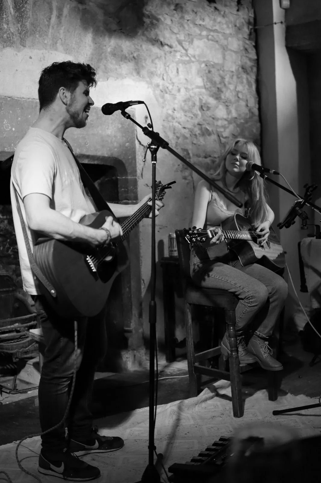 Black and white photo of a young man and woman playing guitars and singing in a cozy, rustic setting with a stone wall background.