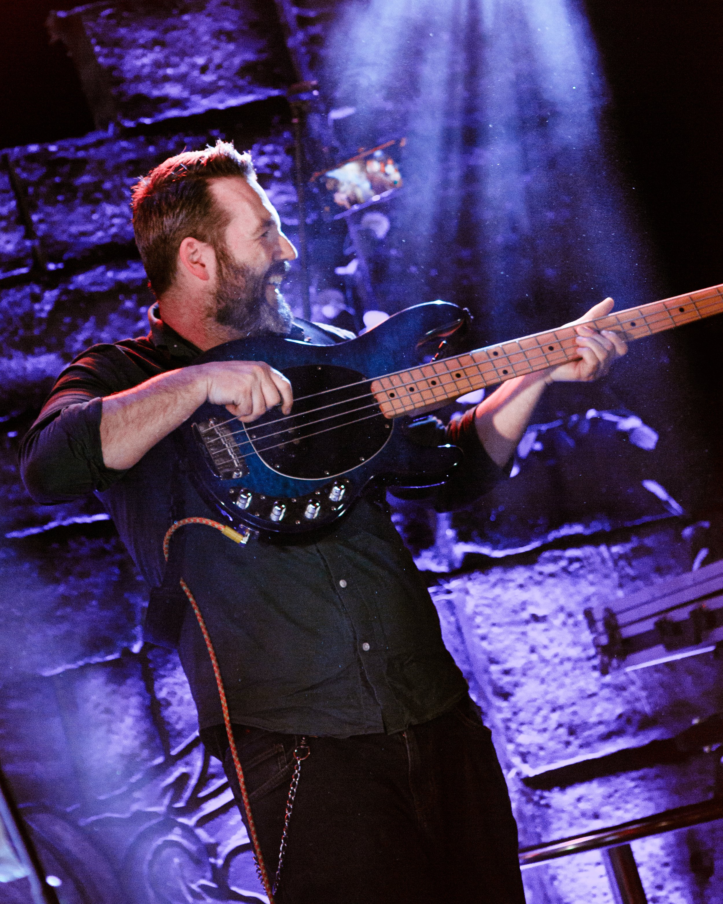 A man with a beard playing an electric bass guitar, smiling, in a setting with blue and purple lighting and a brick wall background.