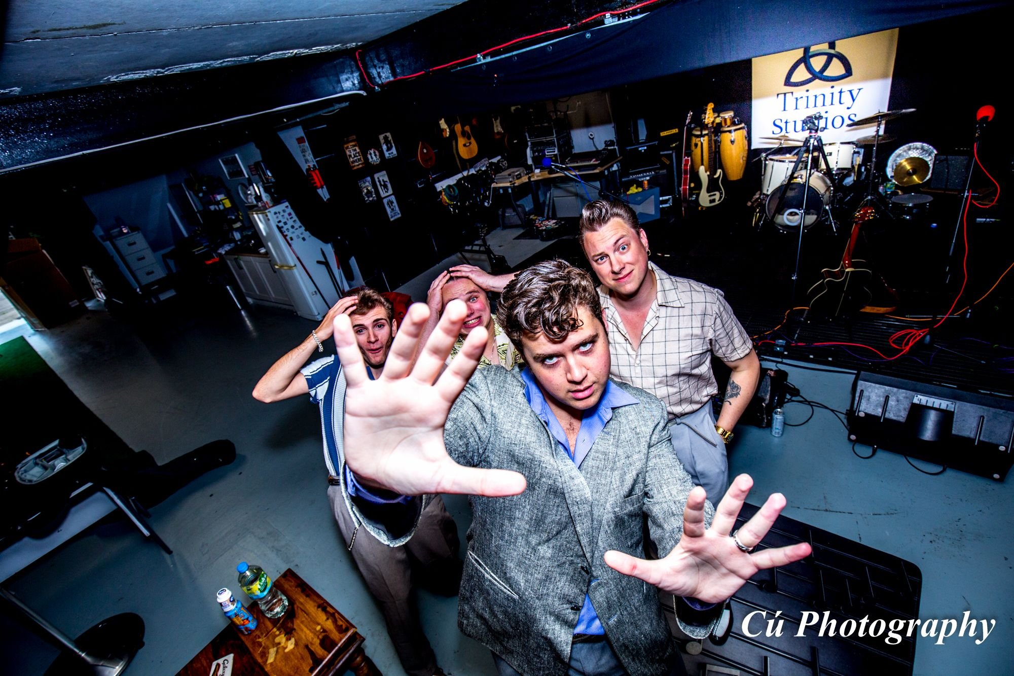 Four young men posing for a photo in a music studio, with musical instruments like guitars and drums in the background, some posters on the wall, and a Trinity Studios sign. One man reaches out to the camera with his hand.