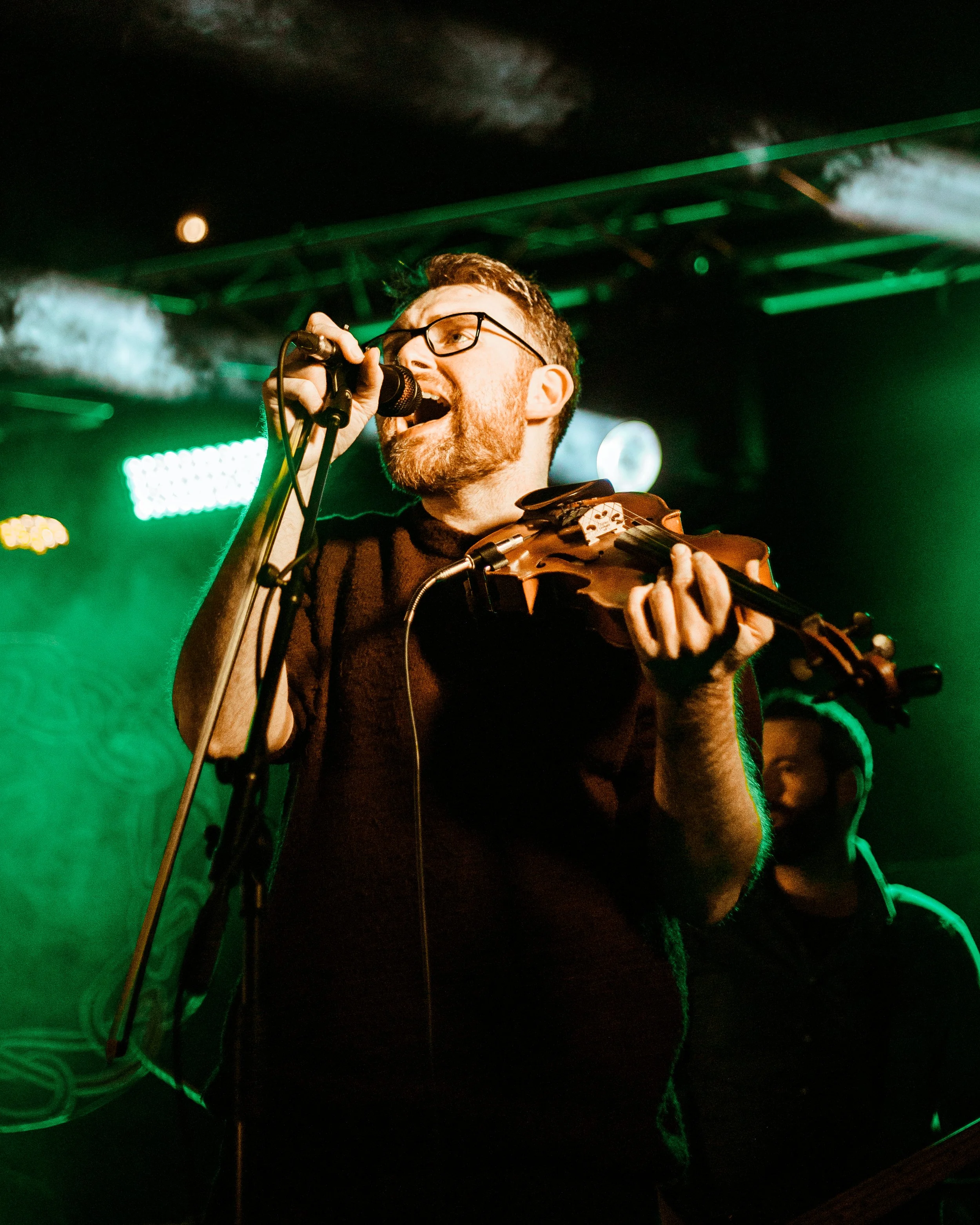 A man with glasses and a beard singing into a microphone while playing a violin on stage with green lighting.