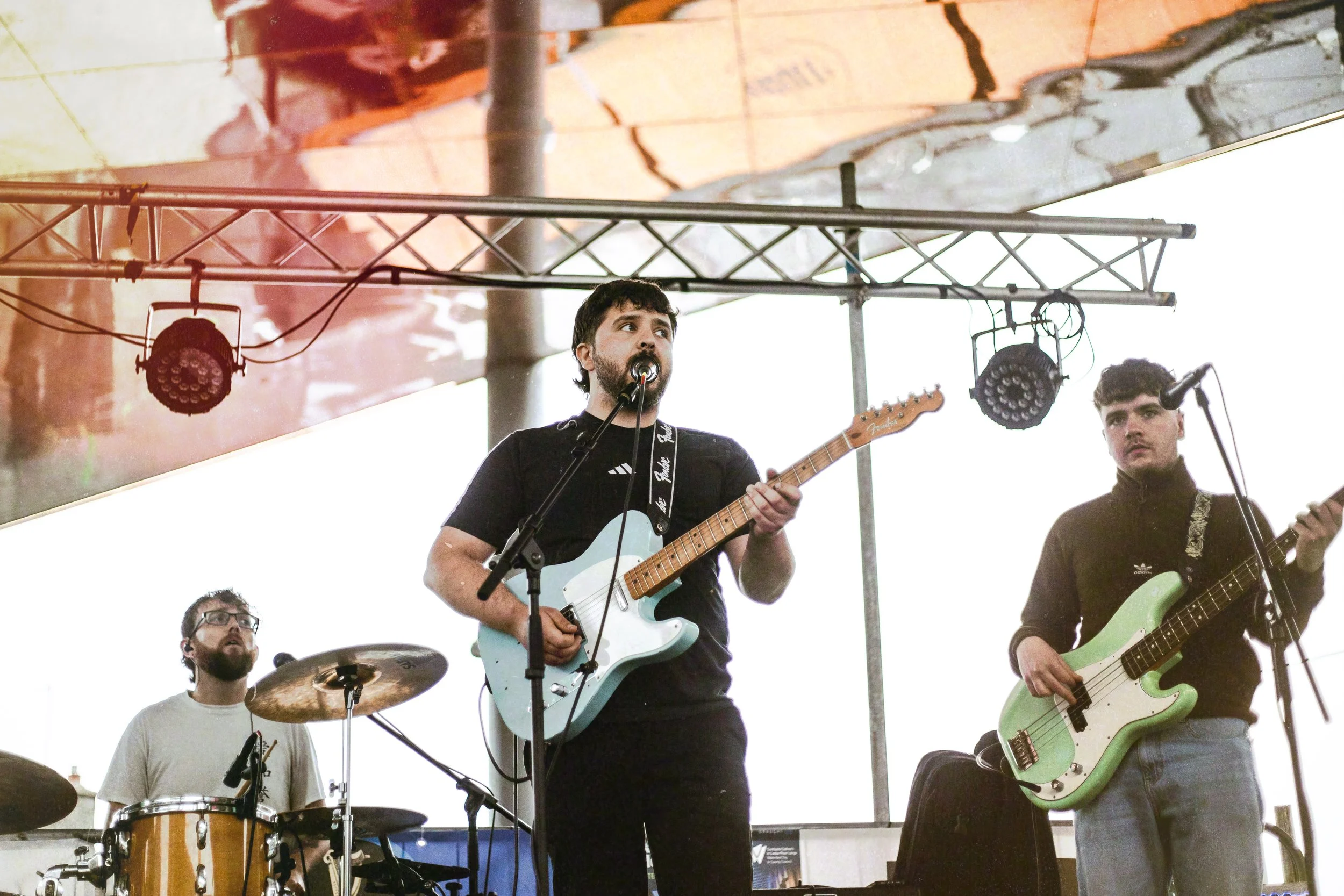 A band performing on stage with four members visible. The lead singer and guitarist in the center has dark curly hair and a beard, playing a light blue electric guitar and singing into a microphone. To his right, a bass guitarist with dark hair, wear