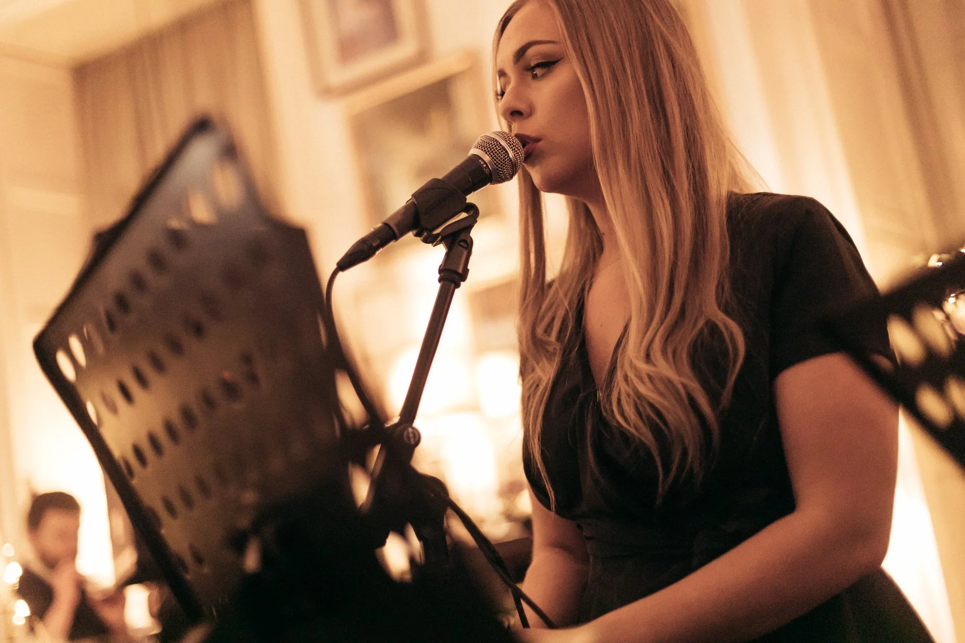 A woman with long wavy blonde hair, wearing dark eye makeup and a black outfit, is singing into a microphone while reading sheet music on a stand. The background is softly lit with warm tones, suggesting an indoor performance or rehearsal setting.