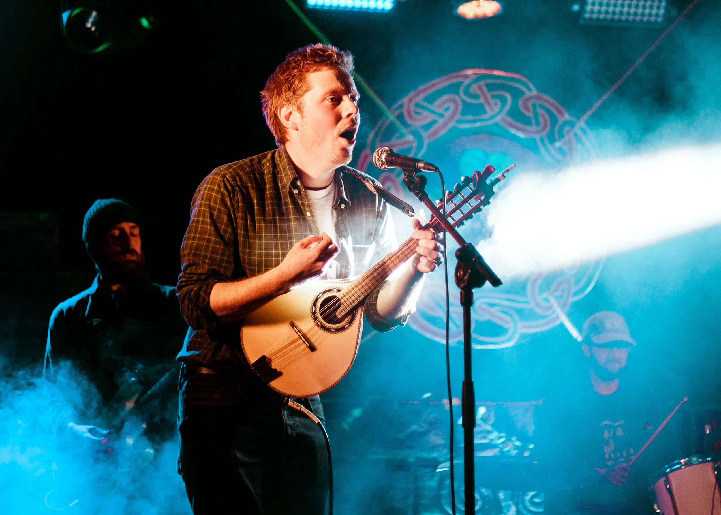 A young man with short hair singing into a microphone while playing an acoustic guitar on stage, with blue and purple lighting and a decorative background.