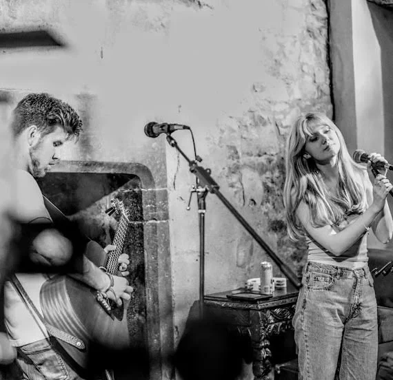 Two young women performing music on a small stage; one playing guitar and the other singing into a microphone.