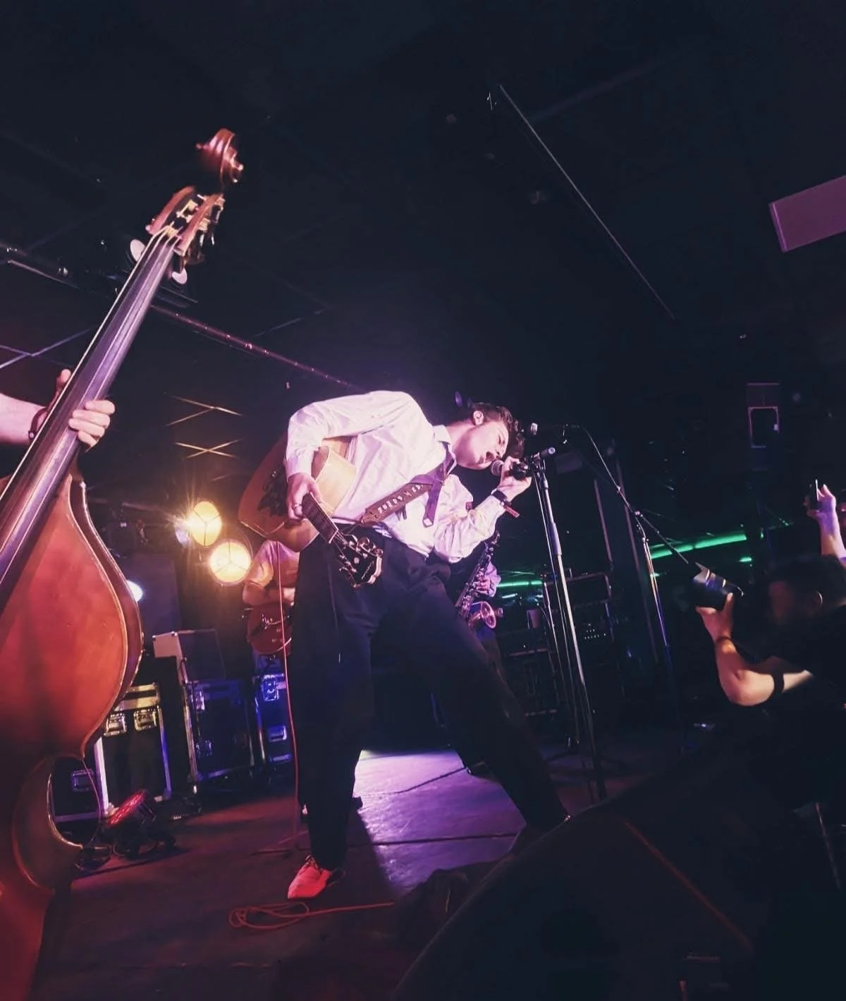 A musician playing a guitar and singing into a microphone on stage, with a bassist playing a double bass and a photographer taking pictures in a dimly lit venue with stage lighting.