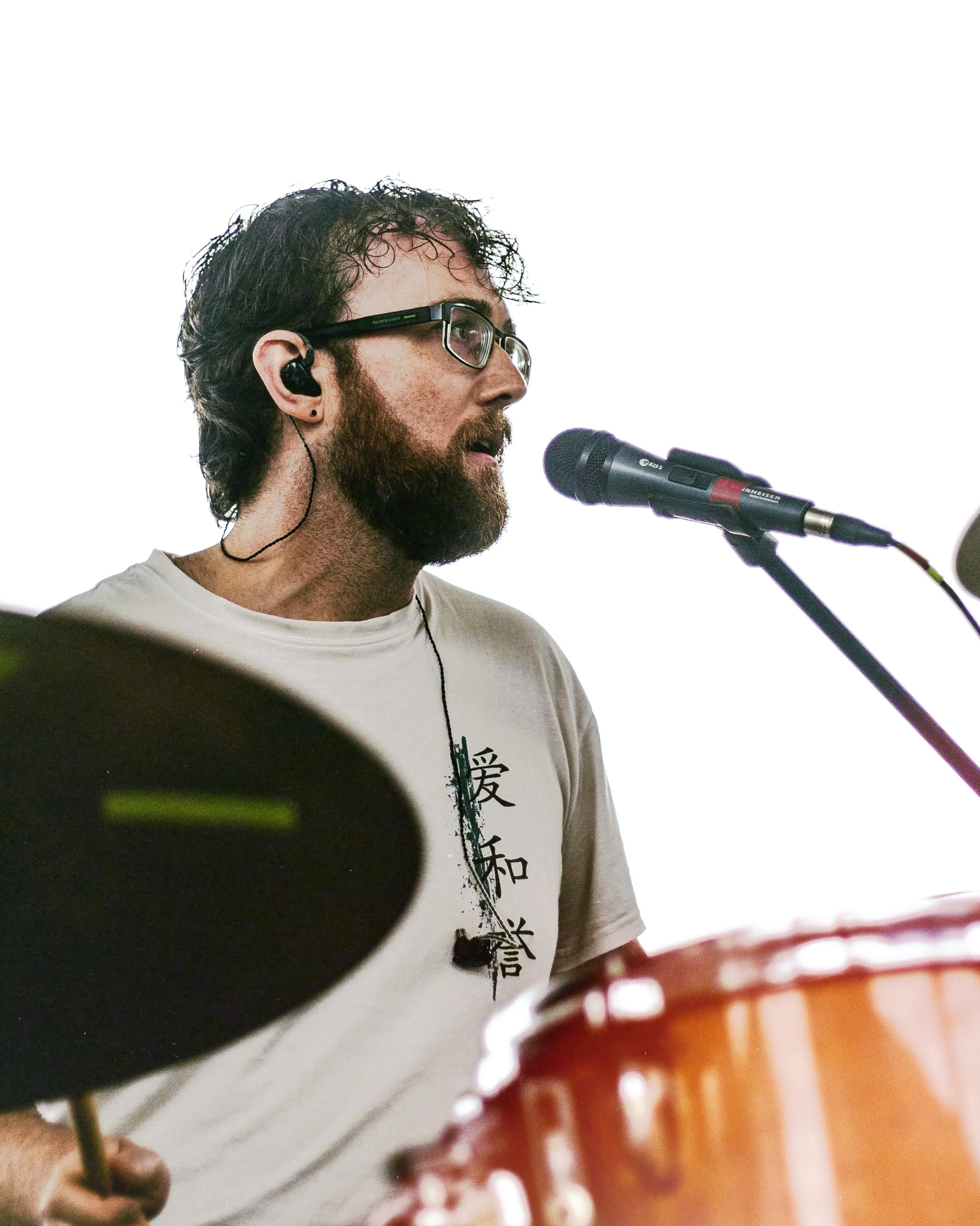 A man with glasses and a beard speaking into a microphone, wearing a white T-shirt with Japanese characters on it, and black in-ear headphones.