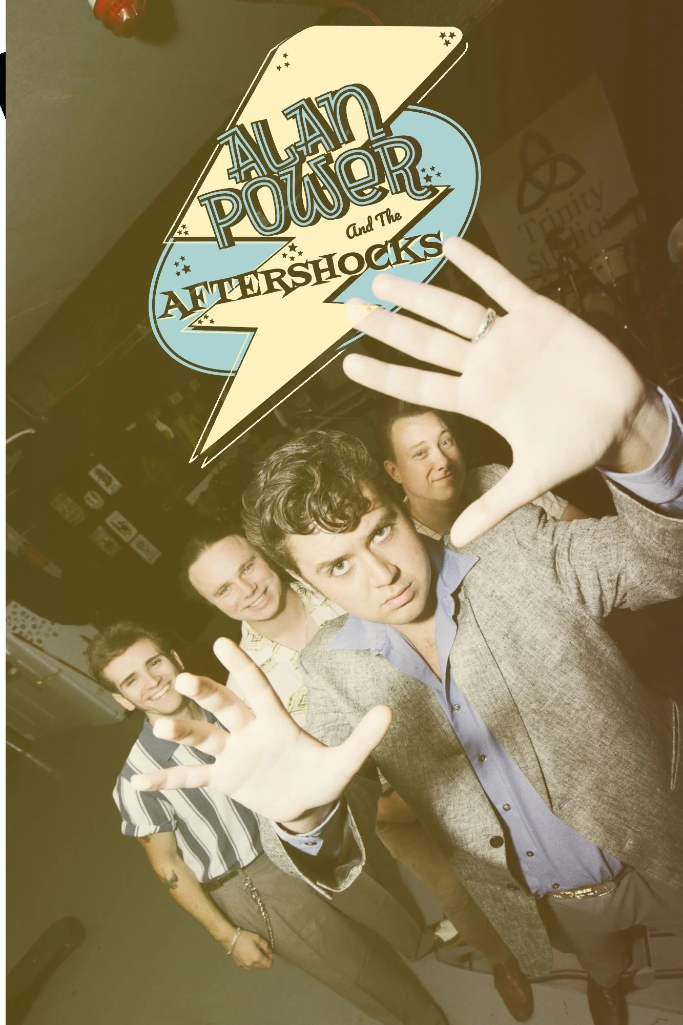 A group of young people, some smiling and some with serious expressions, posing in front of a promotional logo for a band called 'Alan Power and The Aftershocks.' The logo appears as a large cartoon lightning bolt with text over it.