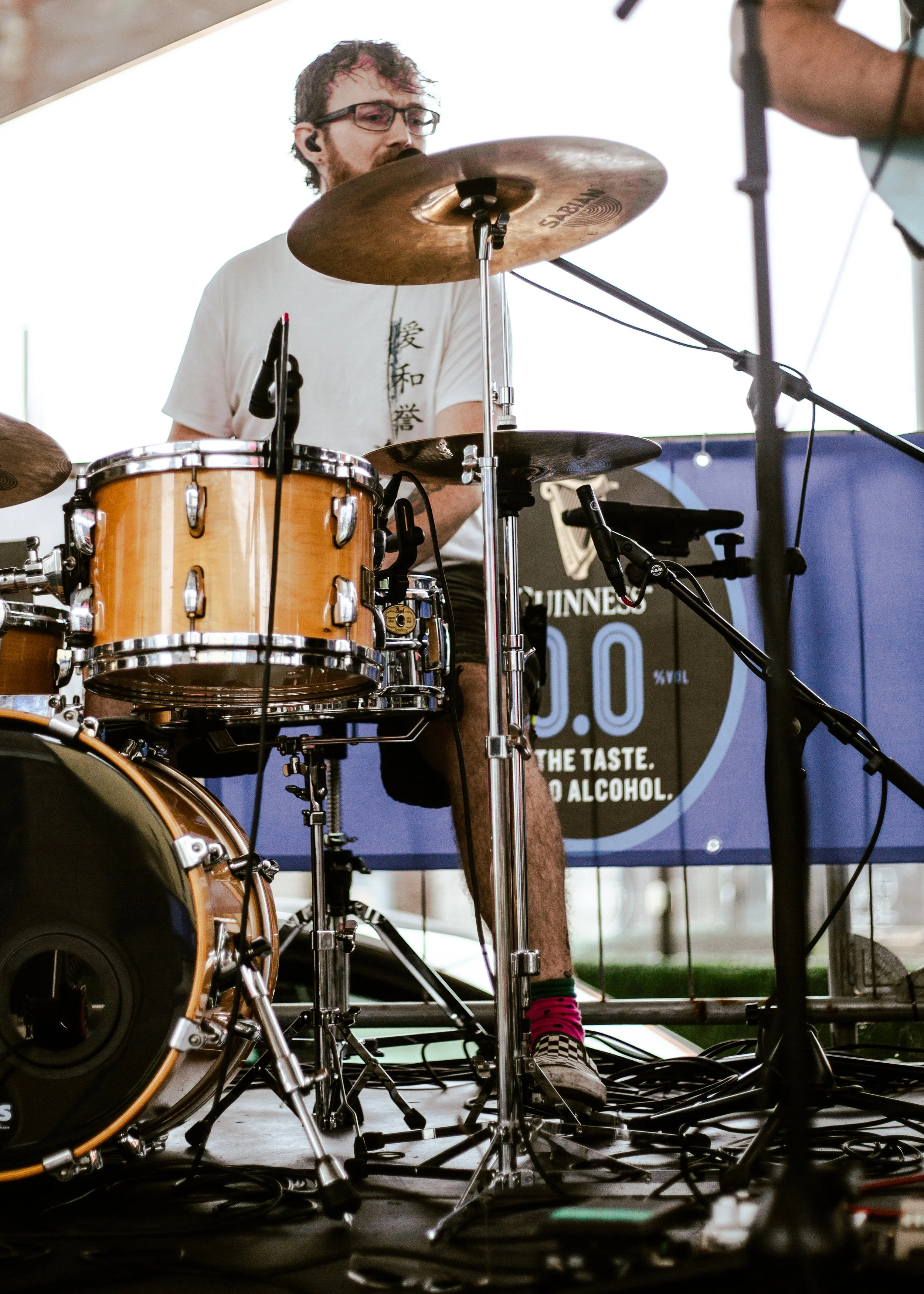 A musician playing drums on a stage, with focus on the drum kit and various equipment wires on the floor. The musician has dark, curly hair, glasses, and is wearing a white T-shirt with Asian characters, pink and black checkered shoes, and socks.