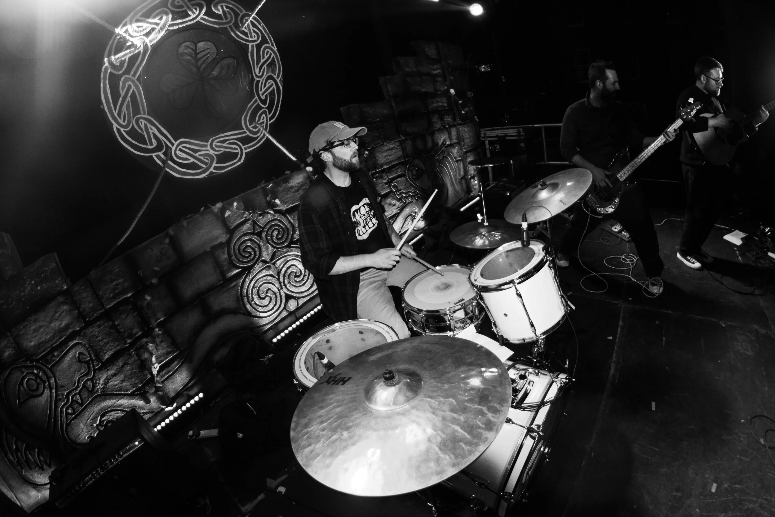 Black and white photo of a band performing on stage. A drummer with glasses, a hat, and a beard is playing drums, accompanied by a bassist and a guitarist. There is a large Celtic knot design with a four-leaf clover in the center hanging on the wall behind them.