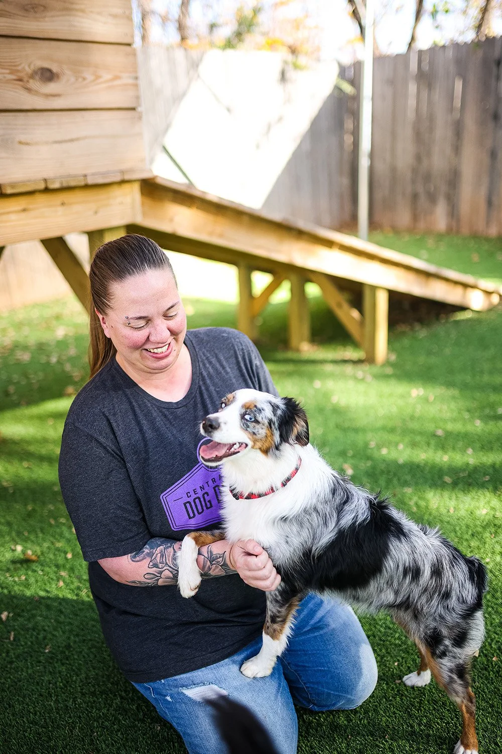 A woman with tattoos on her arm is smiling and holding an Australian Shepherd dog with a merle coat pattern, outside in a backyard with green grass and a wooden dog agility ramp in the background.