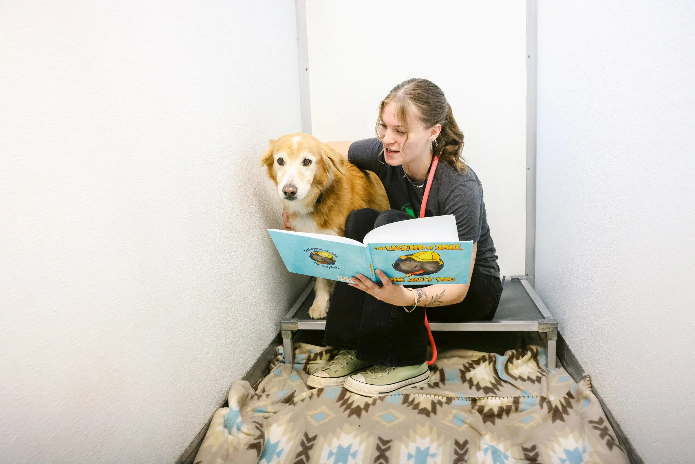 Young woman reading a children's book titled 'The Legend of Jake, the Salty Dog' to her dog in a small, enclosed space with white walls, sitting on a patterned blanket on the floor.