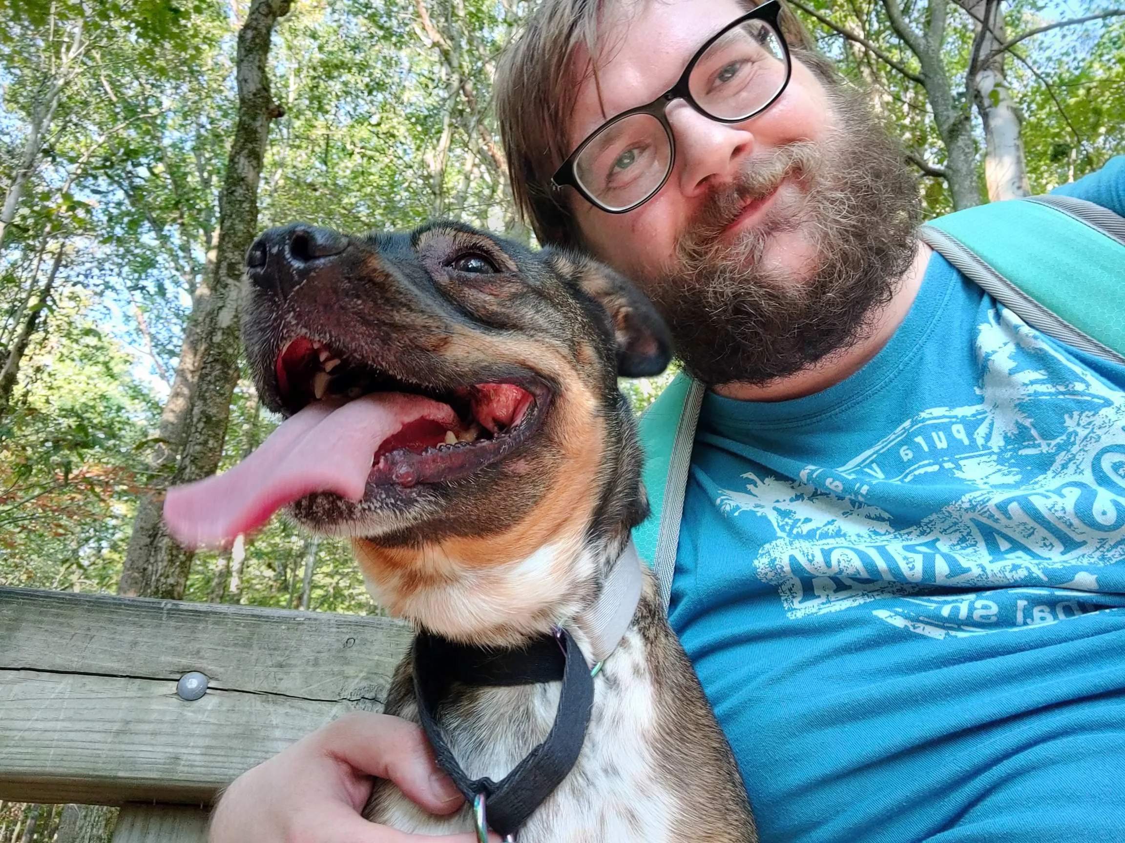 Trevor Schwall and Penny taking a selfie with a smiling dog with its tongue out, outdoors in a wooded area.
