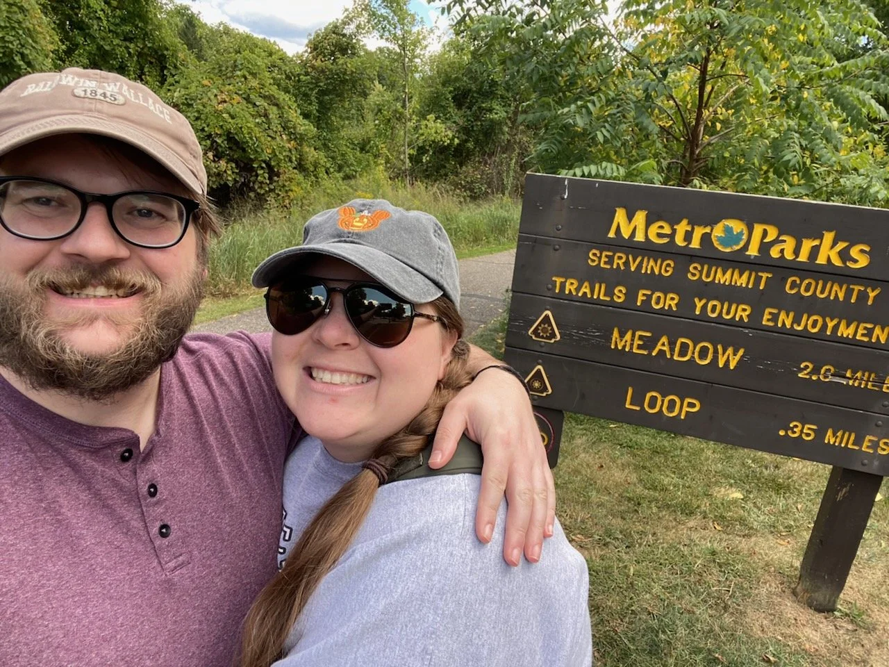 Trevor Schwall and his partner near a trail sign for Metro Parks, serving Summit County, with green trees and shrubs in the background.