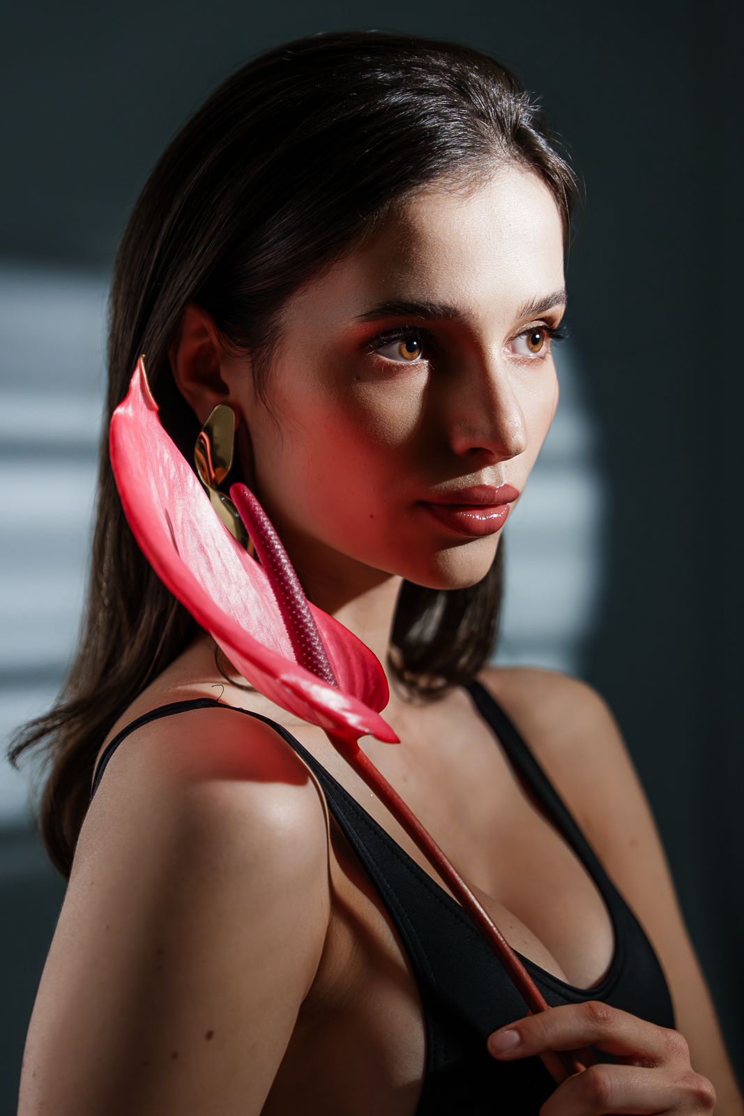 A woman with dark brown hair and wearing a black tank top holding a pink and red anthurium flower close to her face.