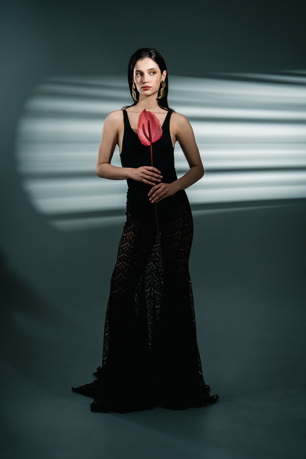 A woman in a black dress holding a red flower-shaped prop indoors with striped light shadows on the wall behind her.