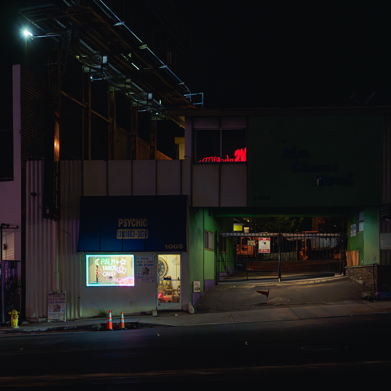 Nighttime street scene with a building that has a neon sign for palm tarot cards reading, an illuminated psychic sign, and some traffic cones outside. The building has a covered entrance and a parking lot behind a gate.