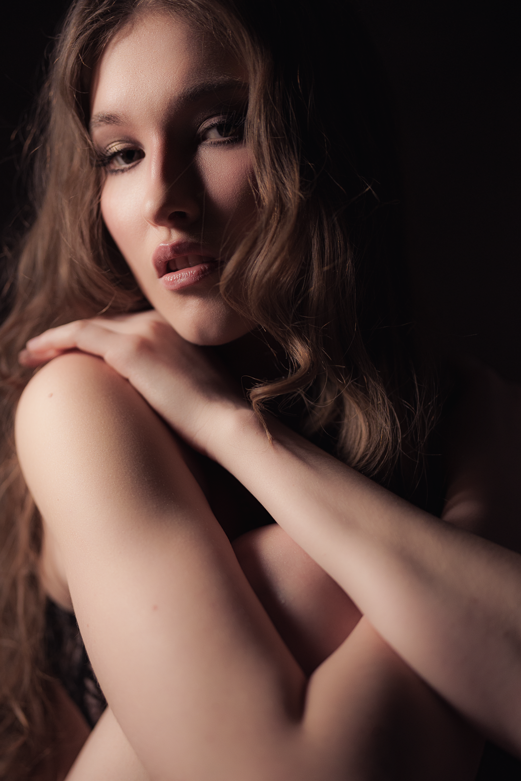 A young woman with wavy brown hair posing with her hand on her shoulder in a dark setting.