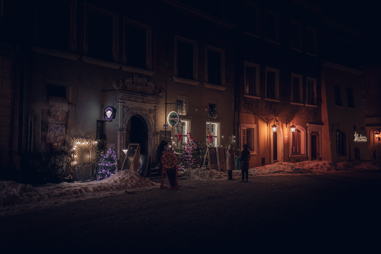 Nighttime street scene with illuminated restaurant signs, decorated Christmas trees, and snow on the ground, with two people standing outside in conversation.