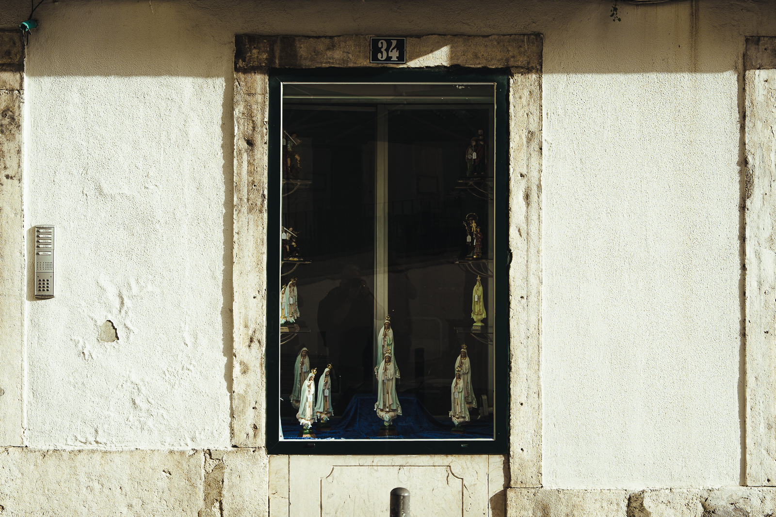 A window display with religious figurines, featuring several statues of saints, set inside a building with off-white textured walls. The building has the number 34 above the window.