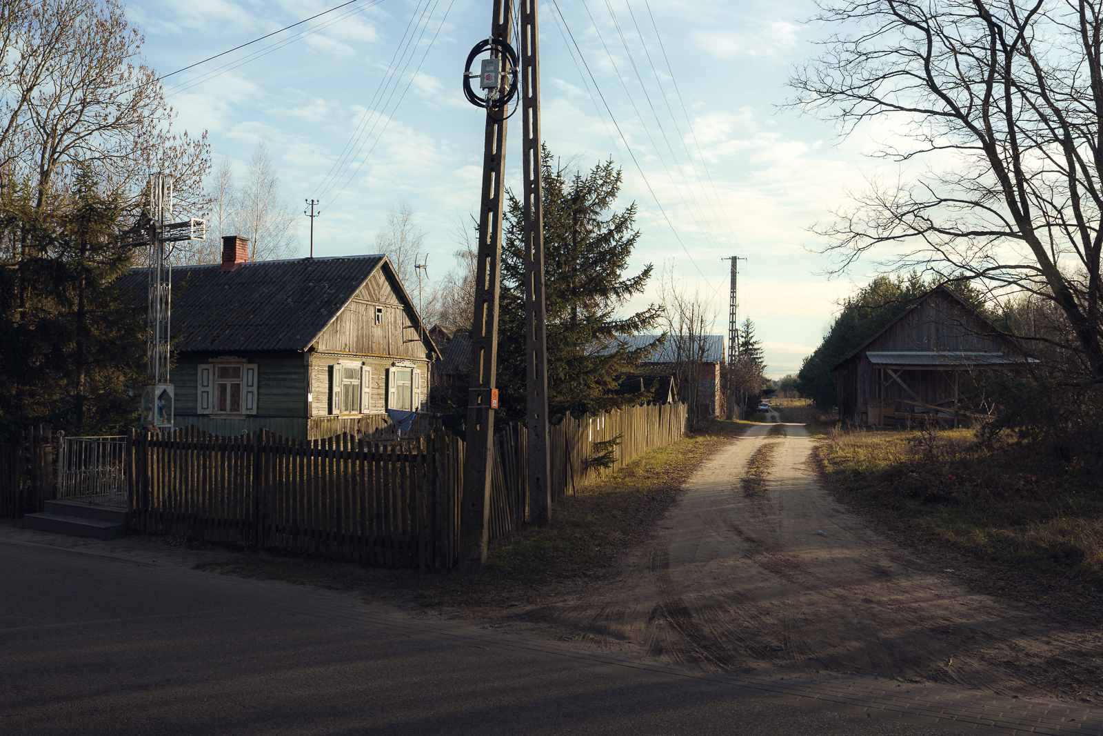 A rural dirt road crossing a paved road with houses and trees on both sides and utility poles under a partly cloudy sky in a countryside setting.
