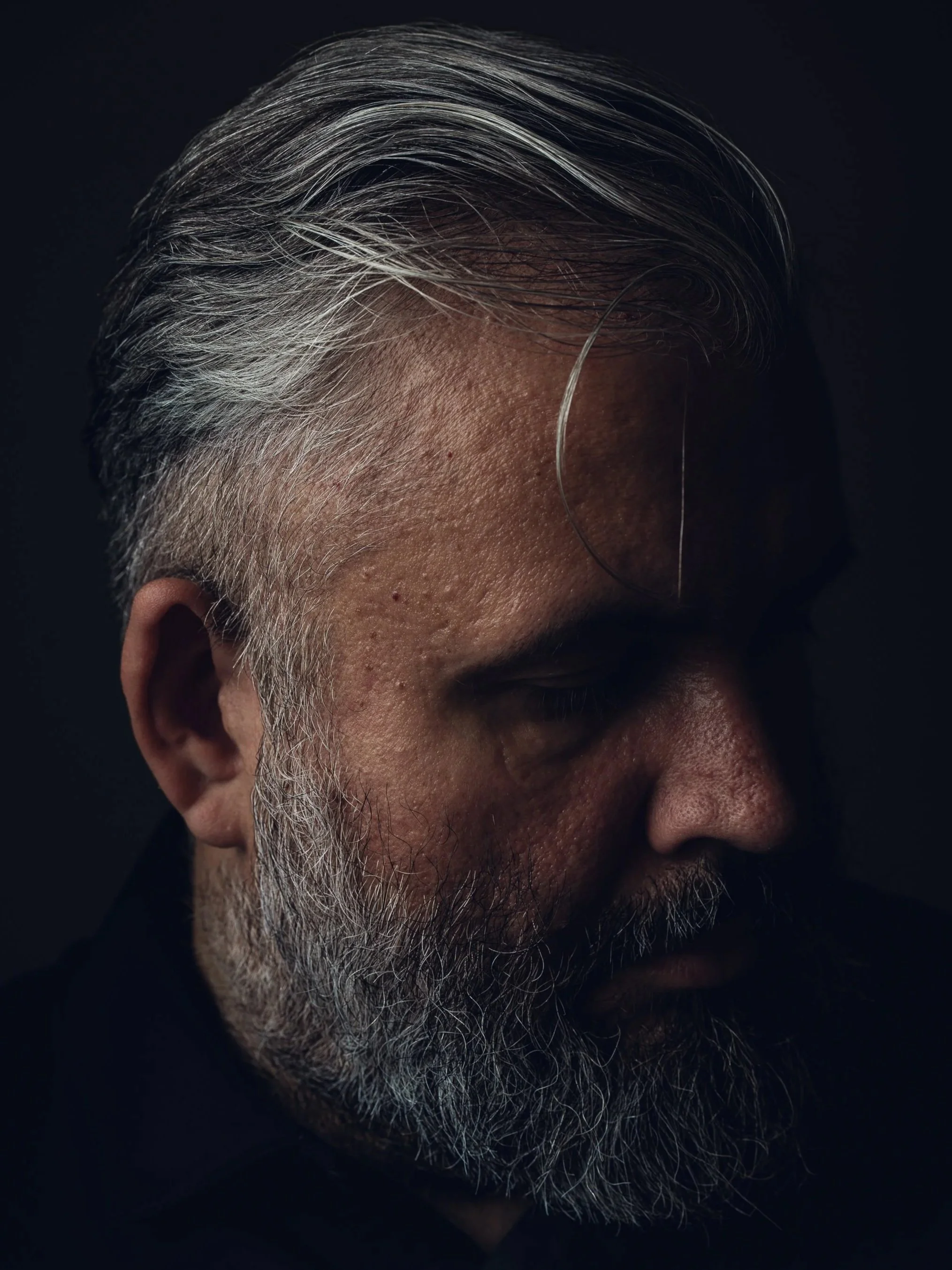 Side profile of a man with gray hair and beard, looking down, against dark background.