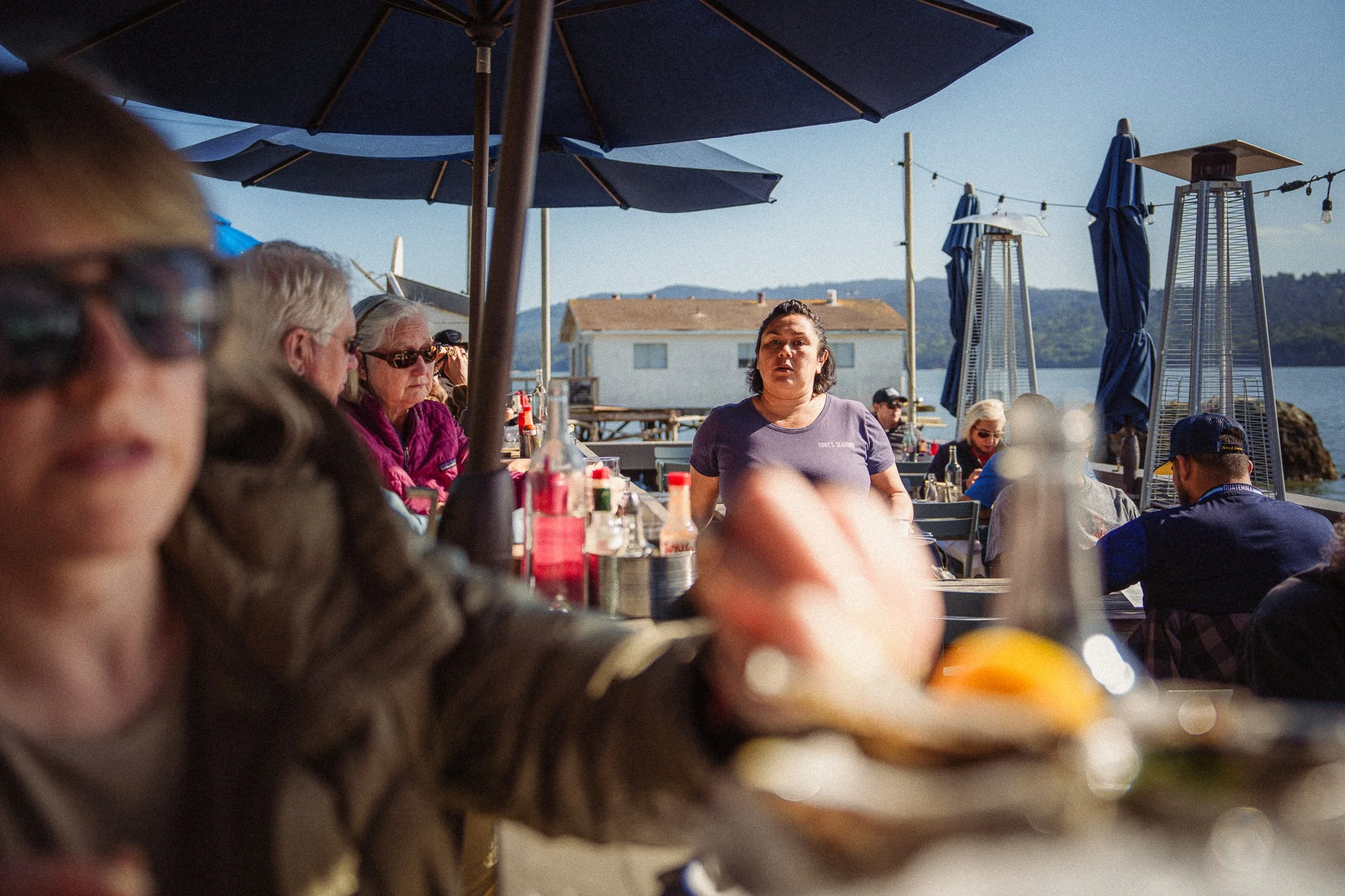 People dining outdoors at a waterfront restaurant with umbrellas, heaters, and scenic lake view in the background.