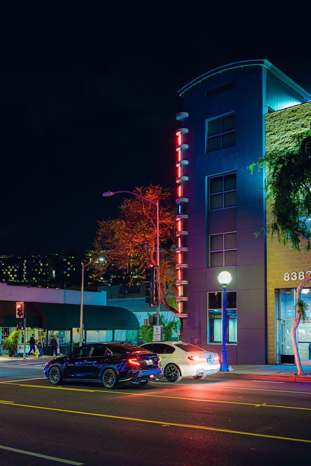 Nighttime street scene with colorful neon lights on a building, two cars parked, and a few pedestrians crossing the street.