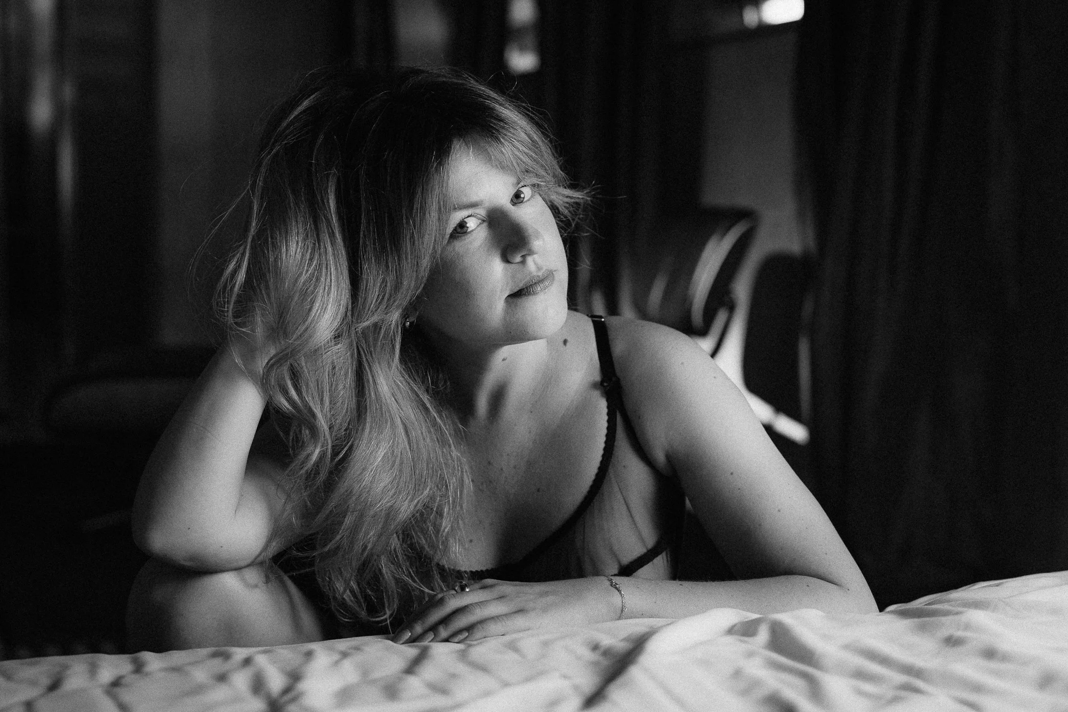 A black-and-white photo of a young woman with wavy hair, lying on a bed with her head supported by her hand, looking at the camera with a soft expression.