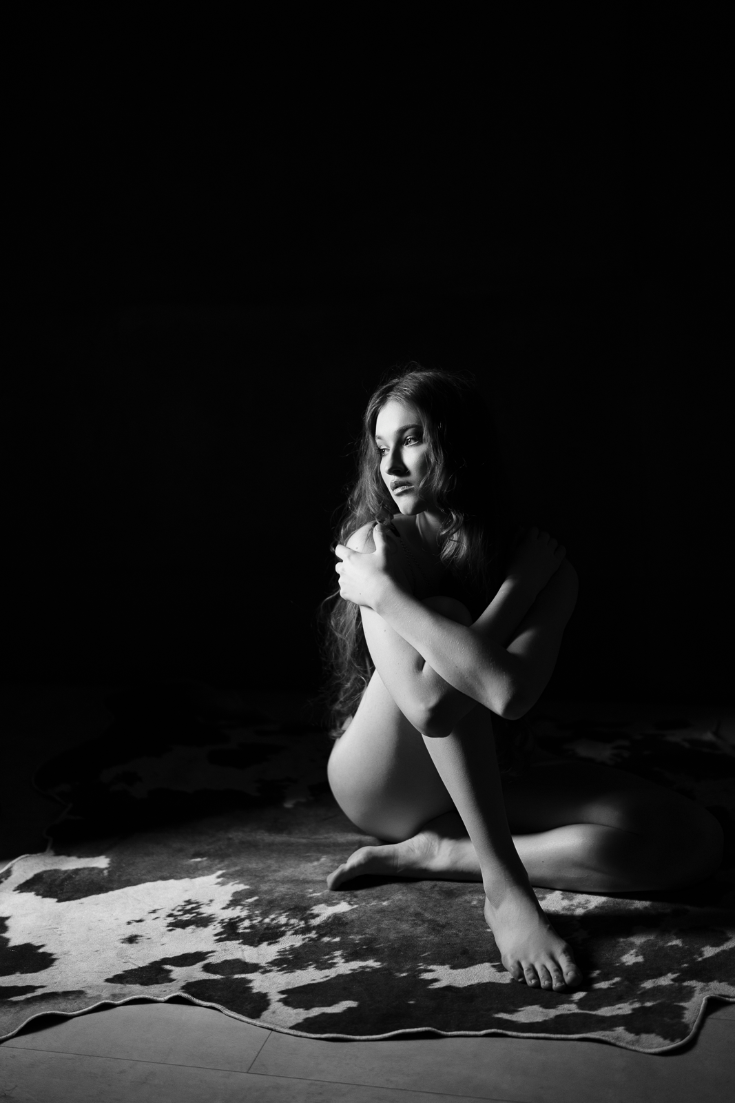 Black and white photo of a young woman with long wavy hair, sitting on a cowhide rug on the floor, with a dark background, gazing thoughtfully to her right, hugging her knees.