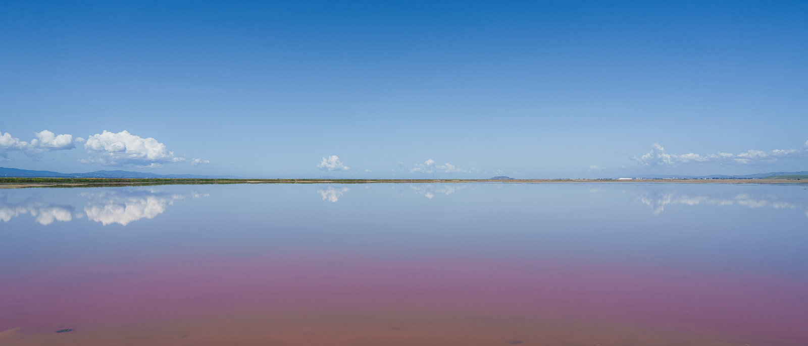 A wide view of a flat landscape with a calm, reflecting body of water under a blue sky with scattered clouds, and distant mountains on the horizon.